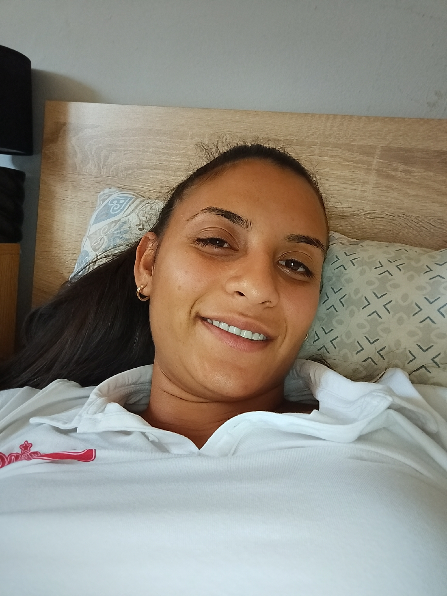 A young woman with dark hair is lying on a bed, looking at the camera. She is wearing a white polo shirt with a red logo on the left side of her chest. She has a gold hoop earring in her left ear. Behind her, there is a wooden headboard and two pillows. One pillow has a blue and white pattern, and the other has a gray and white geometric pattern. The wall behind the headboard is light gray. The scene appears to be indoors, in a bedroom. There is no visible indication of the city of Yambol, Bulgaria.