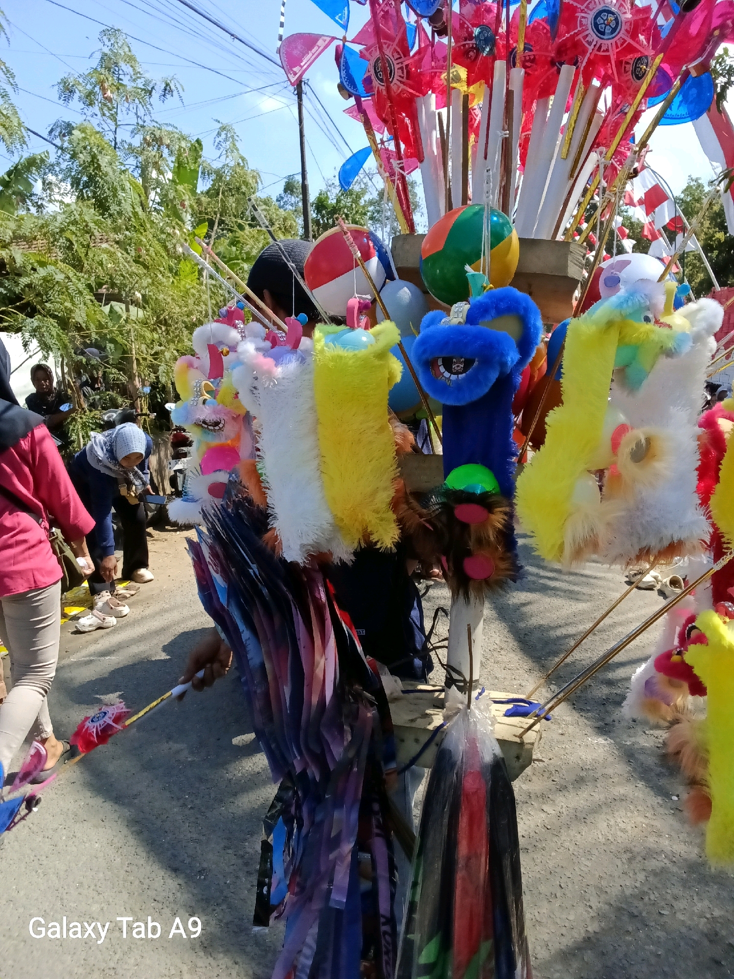 In Bendo, Indonesia, on a sunny day, a street vendor displays an array of colorful toys. In the foreground, various plush toys with bright, fuzzy exteriors in yellow, white, and blue hang from wooden poles. Some of these toys resemble cartoonish animal faces. Further back, a collection of pinwheels with red and blue blades are mounted on poles, along with red and white flags and a striped ball. In the middle ground, several people are interacting. One person wearing a pink jacket and beige pants walks on the asphalt road, while another person wearing a blue hijab and dark clothing bends down, possibly examining or purchasing items. To the left, a person in a black head covering is visible behind a motorcycle. The environment is a paved street with trees and overhead power lines suggesting an outdoor, urban setting.