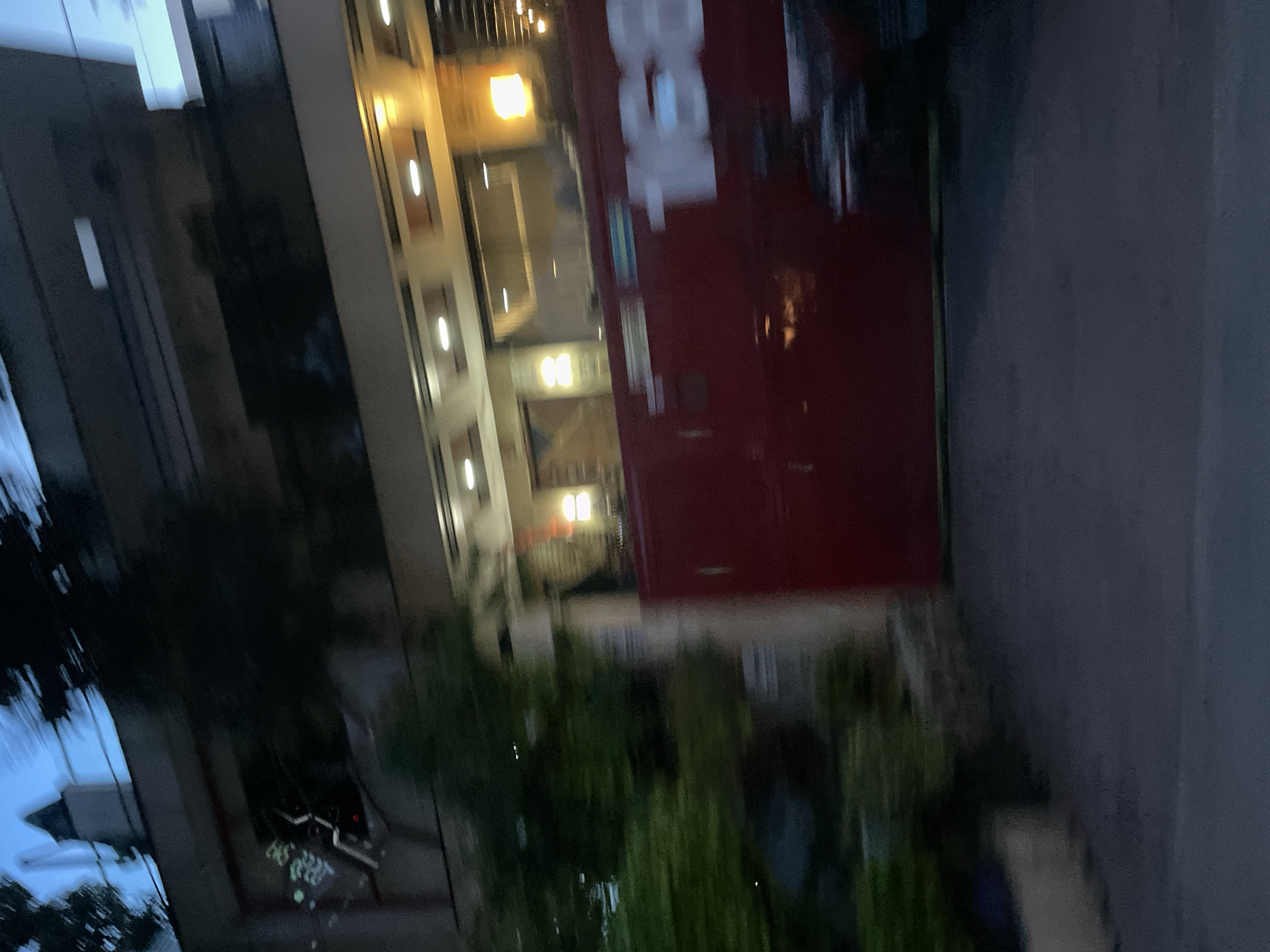 This media file depicts a building exterior at dusk, likely in Davao City, Philippines. The building features a prominent red wall with white lettering that appears to spell "BGC". To the left of the red wall, there's a section of the building with multiple floors, illuminated by warm, yellow lights. The architecture suggests an urban commercial or residential setting.
In the foreground, there are lush green plants, possibly landscaping, creating a natural element against the man-made structures. The lighting indicates it is either early evening or late afternoon, with the sky visible as a deep blue in the reflection on the building's surfaces.
There are some faint reflections on the building's facade, including what might be streetlights or other nearby structures. A digital display is visible in one of the reflections, showing a series of numbers that are too indistinct to read clearly, but it may be a clock or date.
The overall impression is that of a quiet urban scene at the end of the day. No people are clearly visible in the frame. The image is slightly blurred and tilted, giving it a dynamic or candid feel. The presence of the "BGC" text could suggest a business or commercial area, though it's not definitively identifiable without further context.