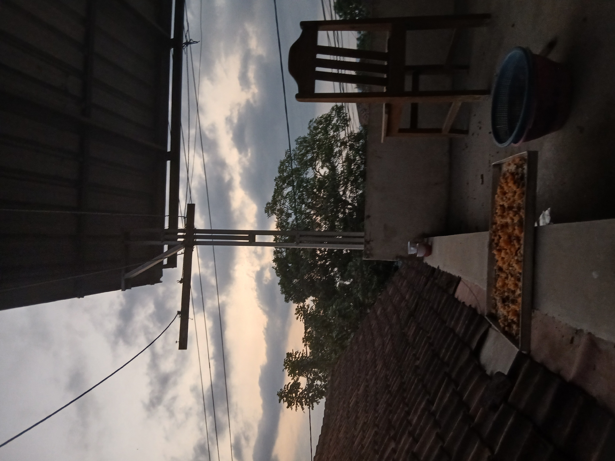 A low-angle, vertical outdoor shot depicts a rooftop scene at dusk in Tenggarang, Indonesia. The foreground features a metal tray filled with what appears to be dried food items, possibly grains or small pieces of fruit, positioned on a concrete ledge. To the right of the tray, a blue plastic colander rests inside a larger red bowl. Further back, a wooden chair with a slatted back and seat stands on the concrete floor. The background reveals a tiled roof on the left, a wall and trees in the middle ground, and a cloudy sky with soft hues of orange and yellow from the setting sun. Overhead, several electrical wires crisscross the frame, supported by metal poles. The overall lighting suggests twilight.