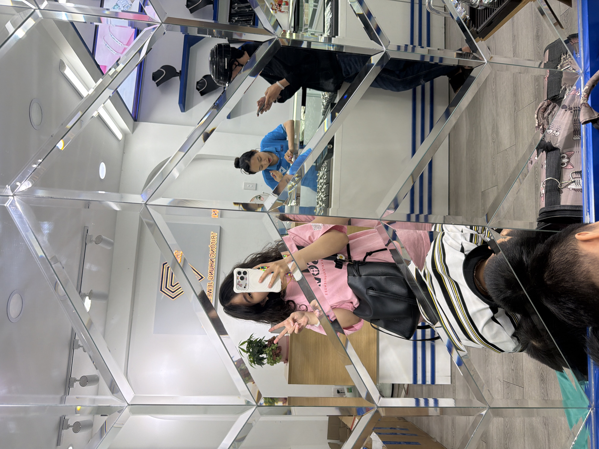 This image captures a vibrant interior scene, likely a retail store in Đà Nẵng, Vietnam, characterized by walls and surfaces covered in a mosaic of angled mirrors, creating multiple reflections and a fragmented view. The primary subject is a young woman, seen in a prominent reflection, taking a selfie with a white smartphone. She wears a pink t-shirt and has a black backpack on her shoulder, making a peace sign with her left hand. Her long, dark hair frames her face.
Numerous reflections showcase different parts of the store and its occupants. In the background, a man wearing a black helmet and jacket is visible, looking at his phone. Another reflection shows a woman in a blue uniform shirt, possibly a store employee, working at a counter. Other details caught in the reflections include overhead lighting fixtures, a light-colored wooden floor, a wall with blue vertical stripes, and a small potted plant. Part of a bag or case with cat patterns is also visible.
A prominent sign, reflected several times, features a golden, geometric, stylized 'D' logo. The actual sign, partially obscured behind the selfie-taker, appears to read "THE DIAMOND KINGDOM". However, in its mirrored reflections, the text distinctly appears as "𝖫𝖵𝖣𝖱𝖤𝖴𝖨𝖲 𝖨𝖭𝖧 𝖯𝖫𝖤𝖬𝖤𝖭𝖳", suggesting a highly stylized font or an optical illusion from the specific mirror arrangement. The overall impression is one of a bright, modern establishment, likely a jewelry or accessories shop, with a playful and immersive design.