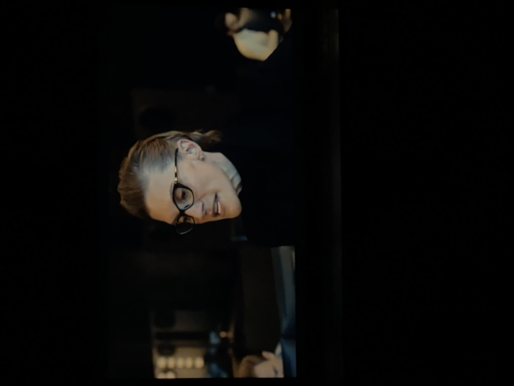 A person is depicted in a dimly lit indoor setting, likely at night or in the evening. The main subject, positioned on the left side of the frame, is a person with light-colored hair, possibly blonde or light brown, pulled back from their face. They are wearing dark-rimmed glasses and looking towards the left, with a slight smile or smirk and their mouth partially open, suggesting they are speaking, laughing, or reacting to something. Their expression conveys amusement or engagement. The background is mostly dark and out of focus, with some blurry, illuminated shapes and another indistinct figure visible further back. The scene suggests a social environment such as a bar, restaurant, or lounge. Based on the provided context, this location could be in Budva, Montenegro. There is no visible text in the image.