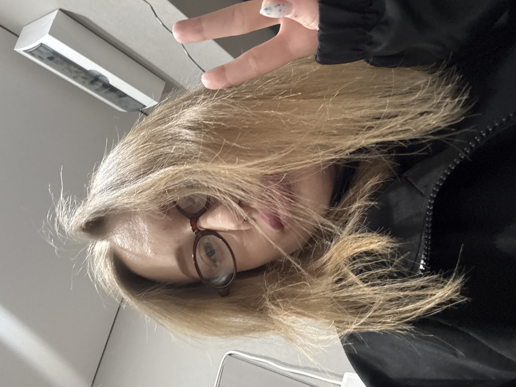 A woman with blond hair, wearing glasses and a black jacket, is holding up two fingers in a "peace" sign. She is indoors, beneath a white, rectangular ceiling light fixture. The location is not evident in this image, and there is no verifiable information indicating it is in Azov, Russia.