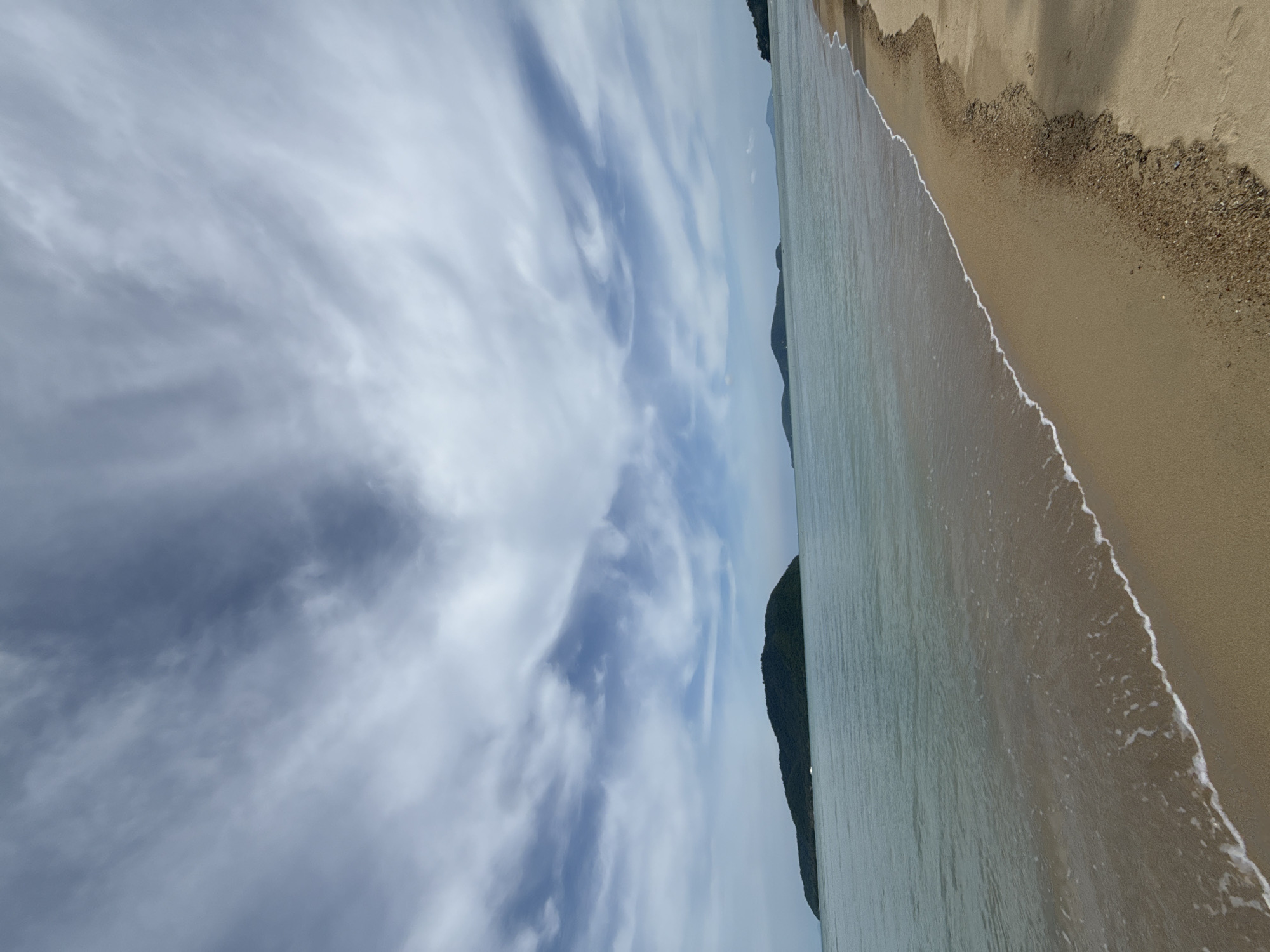 A wide-angle, eye-level view captures a serene beach scene on a cloudy day. The foreground is dominated by a strip of golden sand that slopes gently towards the sea. The sand is coarse, with small pebbles mixed in, and a faint shadow is cast from the right edge of the frame. The water's edge is marked by a thin, foamy white line of gentle waves lapping onto the shore. The sea itself is a tranquil, greenish-blue, extending towards the horizon.
In the middle ground, several islands are visible. The largest, a dark green, forested island, sits prominently to the left of the frame. It appears as a low, elongated shape against the water. Two smaller, more distant islands are visible to the right of the main island, appearing as dark silhouettes against the horizon.
The sky above is a dramatic canvas of white and gray clouds. Wispy, streaky clouds, tinged with darker gray in places, form swirling patterns across the expanse. The overall lighting suggests it is daytime, but the cloud cover creates a diffused, slightly subdued light.
The location is identified as a beach in Rawai, Thailand. The scene conveys a sense of peaceful isolation and natural beauty. There are no people or man-made structures visible in the immediate vicinity.