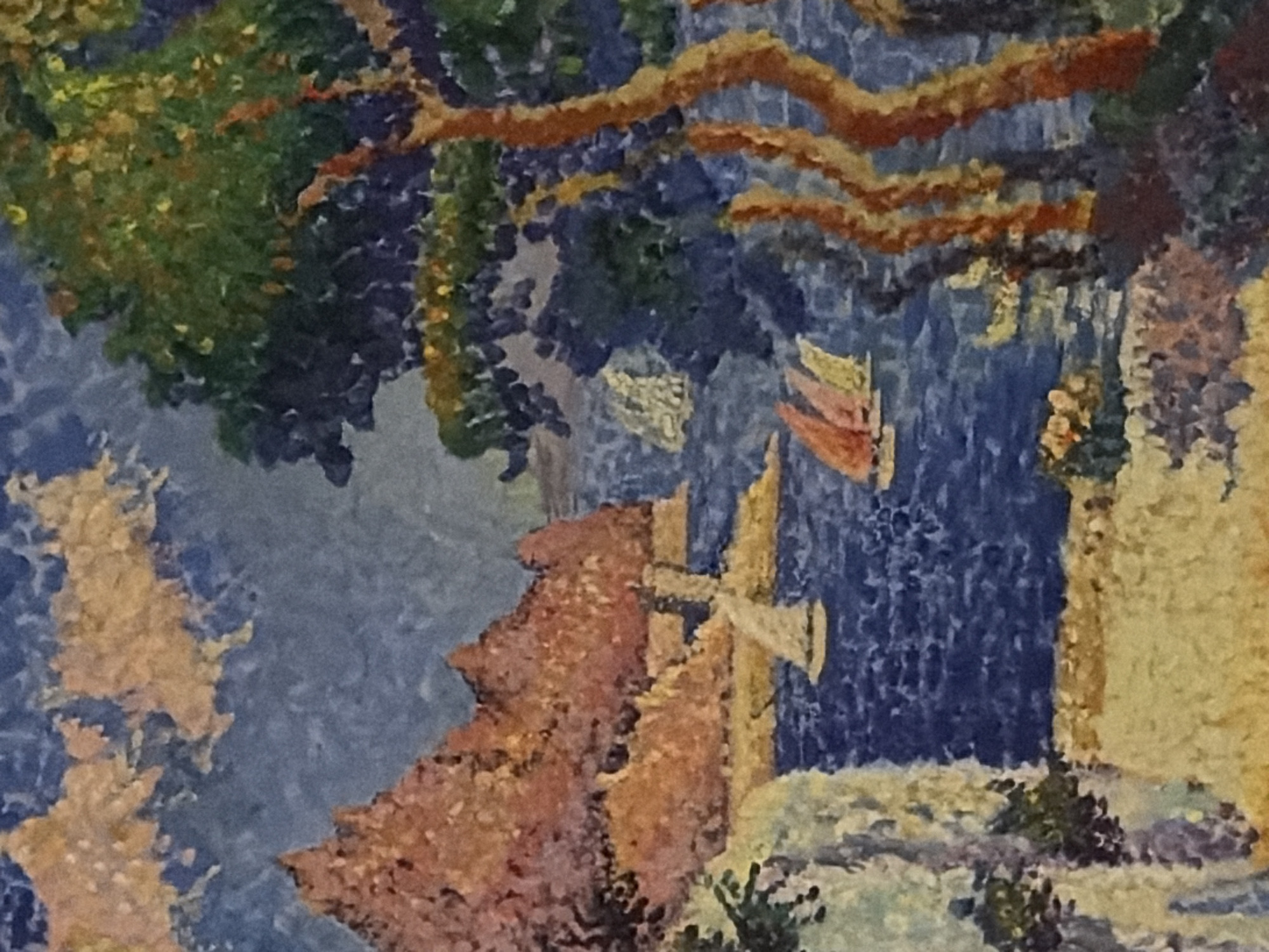 This is a pointillist painting of a coastal scene. In the foreground, to the left, is a light-colored cliff face covered in foliage. To the right of the cliff is a sandy beach with some vegetation. The sea occupies the middle ground, with several sailboats visible. One sailboat is closer to the shore on the left, with white sails. Further out in the sea, three sailboats are clustered together, with sails in shades of red and yellow. In the background, a large, rocky, reddish-brown mountain rises from the sea. To the right of the mountain is a dense green tree with orange-brown branches. The sky above is a mottled blue with light-colored clouds. The painting is rendered with small dots of color. There are no people or any discernible structures associated with the city of Germasogeia Municipality, Cyprus, within the depicted scene.