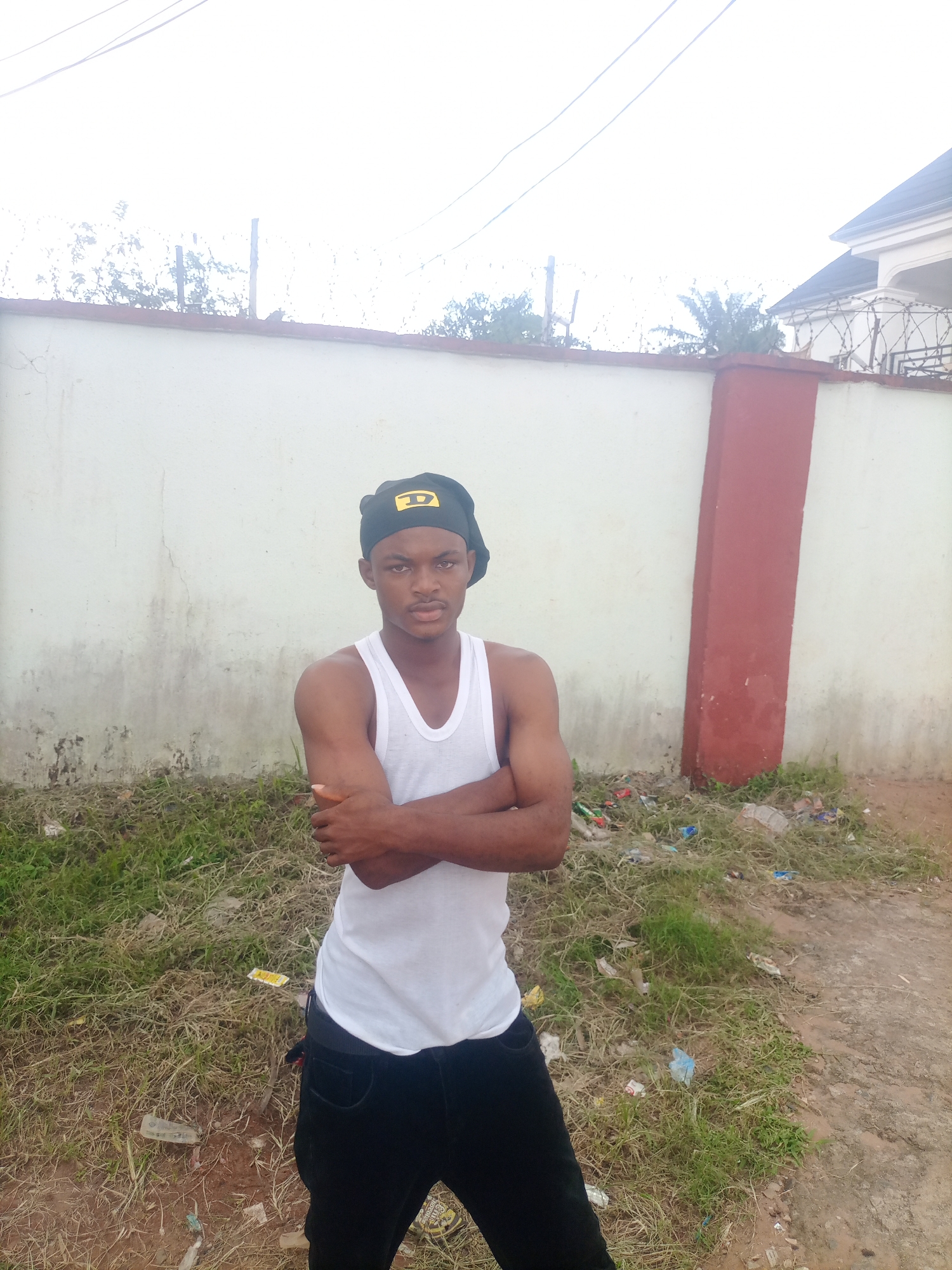 A young Black male stands outdoors, facing the camera with his arms crossed. He is wearing a white tank top, black pants, and a black baseball cap with a yellow logo featuring the letter "D". The background consists of a light-colored wall with some grime and staining, topped with barbed wire. To the right of the wall, a red-painted pillar is visible. The ground in front of the wall is covered in dry grass and scattered litter, including plastic bottles and wrappers. Behind the wall, a glimpse of a building with a dark roof and white facade can be seen. The sky is overcast, suggesting either early morning or a cloudy day. The overall scene appears to be an urban or semi-urban environment in Nkpor, Nigeria, with a slightly unkempt appearance due to the litter. The subject's expression is neutral.