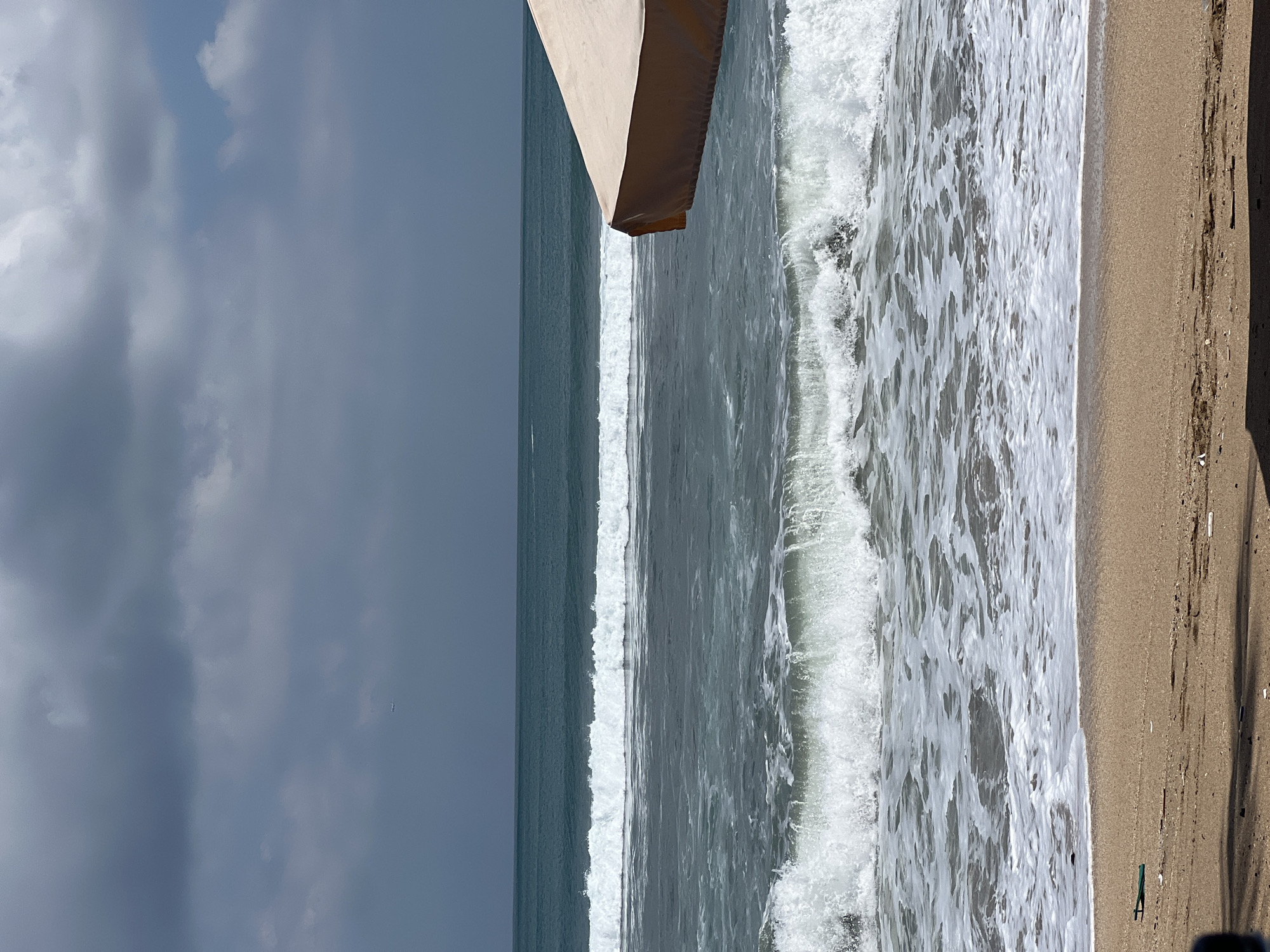 A wide shot captures a serene beach scene in Kuta, Indonesia, under a partly cloudy sky. The foreground shows a sandy beach with scattered debris and a dark shadow on the left. The ocean is calm with gentle waves rolling in, creating white foam as they reach the shore. In the mid-ground, a beige beach umbrella shades a portion of the frame on the right. The background features a vast expanse of ocean merging with a hazy horizon, and a distant airplane is visible in the sky. The overall mood is tranquil and inviting.