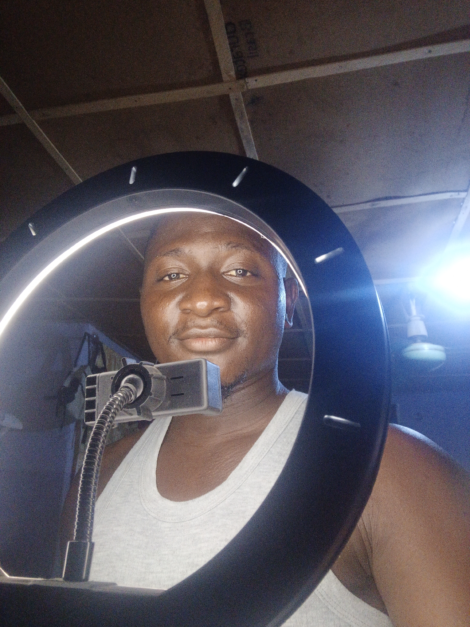 A close-up, medium shot captures a dark-skinned individual from the chest up, framed by a large, illuminated ring light. The person is wearing a white tank top and looking directly at the camera, their face evenly lit by the ring light. A small black device with a flexible metal gooseneck arm is attached to the ring light, positioned in front of the person's chin. The background is dimly lit and shows a wooden ceiling with exposed beams and some electrical wiring. To the right of the ring light, a bright light fixture with a fan-like base is visible. The overall scene appears to be an indoor setting, possibly a room or studio.