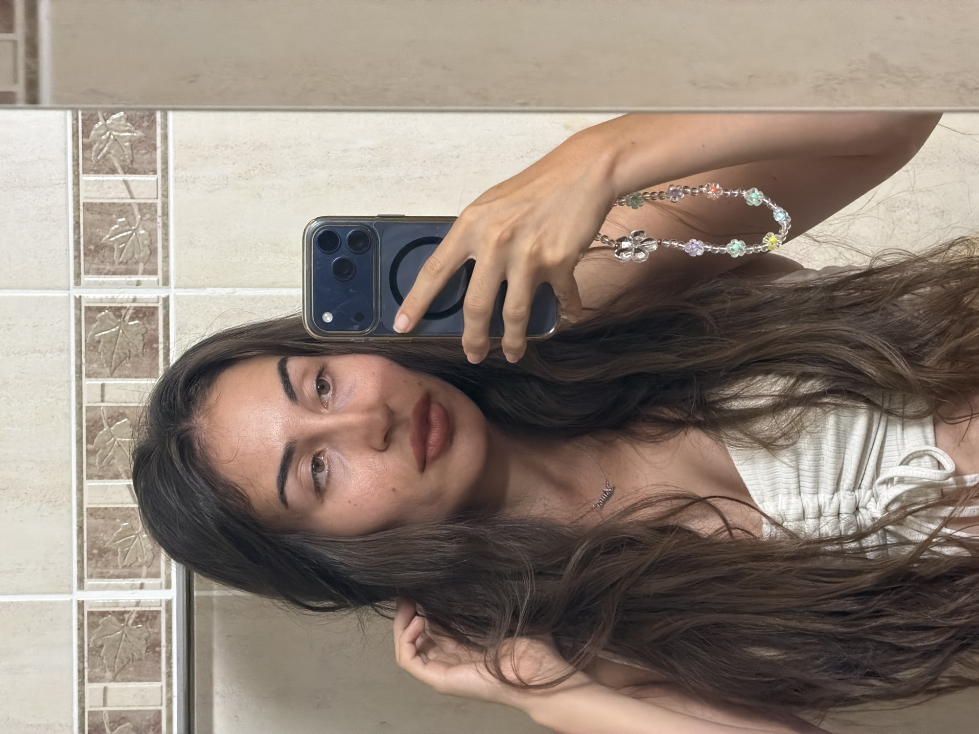 The image is a selfie taken in a bathroom in Tersefanou, Cyprus. The subject is a young woman with long, dark, wavy hair. She is wearing a white, ruched top and a delicate silver necklace with the name "JENNIE" visible. She is holding a smartphone, which is reflecting in the mirror. The phone case has a beaded strap with colorful flower charms. The bathroom wall behind her is tiled with a beige, textured tile and a decorative border with leaf motifs. The lighting appears to be artificial, suggesting it is indoors. The woman's expression is neutral, and she is looking at the camera. The reflection in the mirror shows her arm and the phone she is holding.