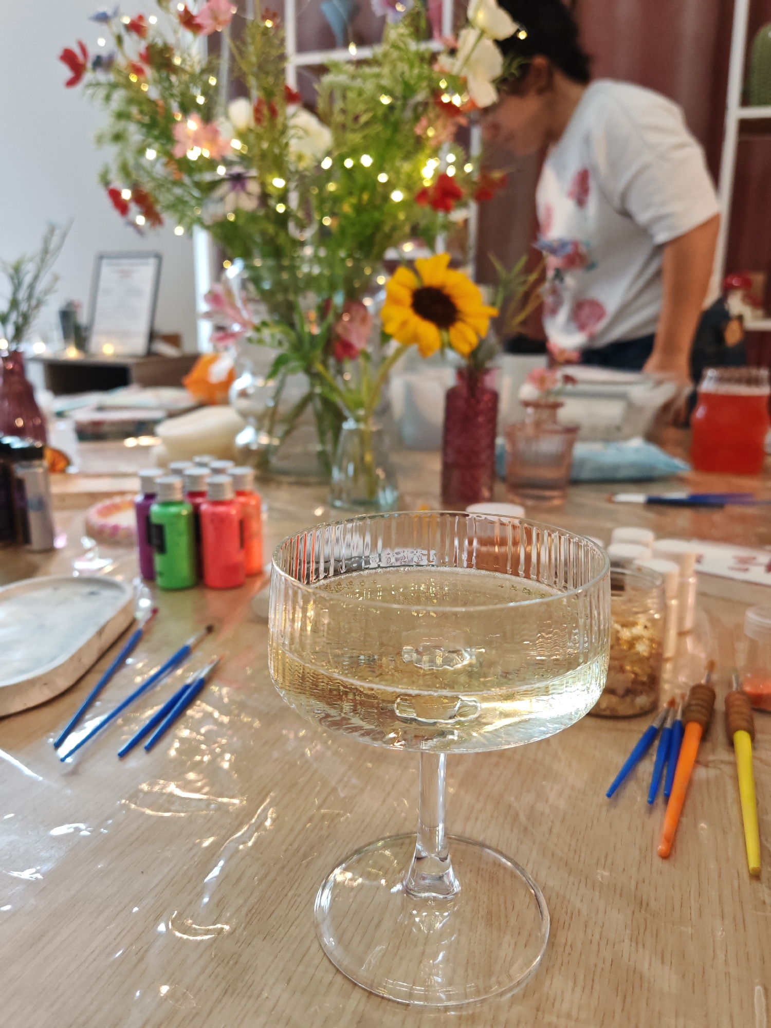The image depicts a close-up view of a table set for a creative activity, likely crafting or painting, with a glass of champagne in the foreground. The table is covered with a clear plastic sheet, revealing a wooden surface beneath. Scattered across the table are various art supplies, including small bottles of colorful paint (purple, green, pink, orange), paintbrushes with blue, orange, and yellow handles, and what appears to be a mold or tray with a grey substance. In the background, a woman is visible, slightly out of focus, wearing a white t-shirt with a floral pattern. She is leaning over the table, suggesting she is engaged in the activity. A large bouquet of wildflowers, some adorned with fairy lights, is prominently displayed in a vase, adding a festive and natural element to the scene. Other decorative elements, such as small glass vases and jars containing glitter or small objects, are also present. The overall atmosphere appears to be relaxed and celebratory, possibly at a workshop, party, or gathering. The image does not contain any specific elements that would allow for localization to Plovdiv, Bulgaria.