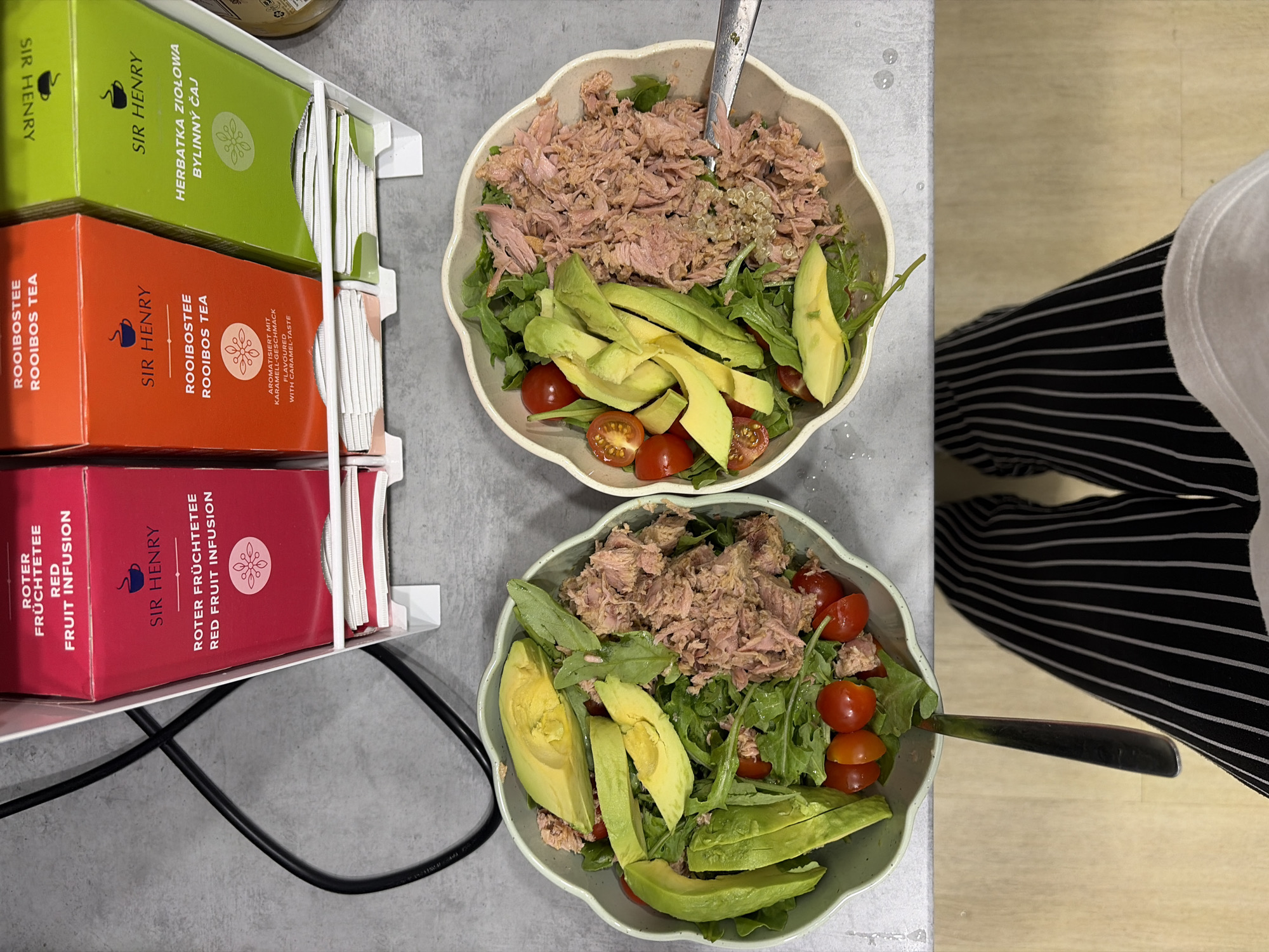 The image shows a close-up, overhead view of two bowls of tuna salad with avocado and cherry tomatoes, placed on a textured gray surface. To the left of the bowls, a black electrical cord is visible. Behind the bowls, a white plastic dispenser holds three boxes of Sir Henry tea: one red box labeled "ROTER FRUCHTETEE RED FRUIT INFUSION," an orange box labeled "ROOIBOSTEE ROOIBOS TEA," and a green box labeled "HERBATKA ZIOLOWA BYLINNY CAY." Each tea box has a small logo of a steaming cup. The bottom portion of the image shows the legs of a person wearing black and white striped pants. The scene appears to be indoors, possibly in a kitchen or dining area.