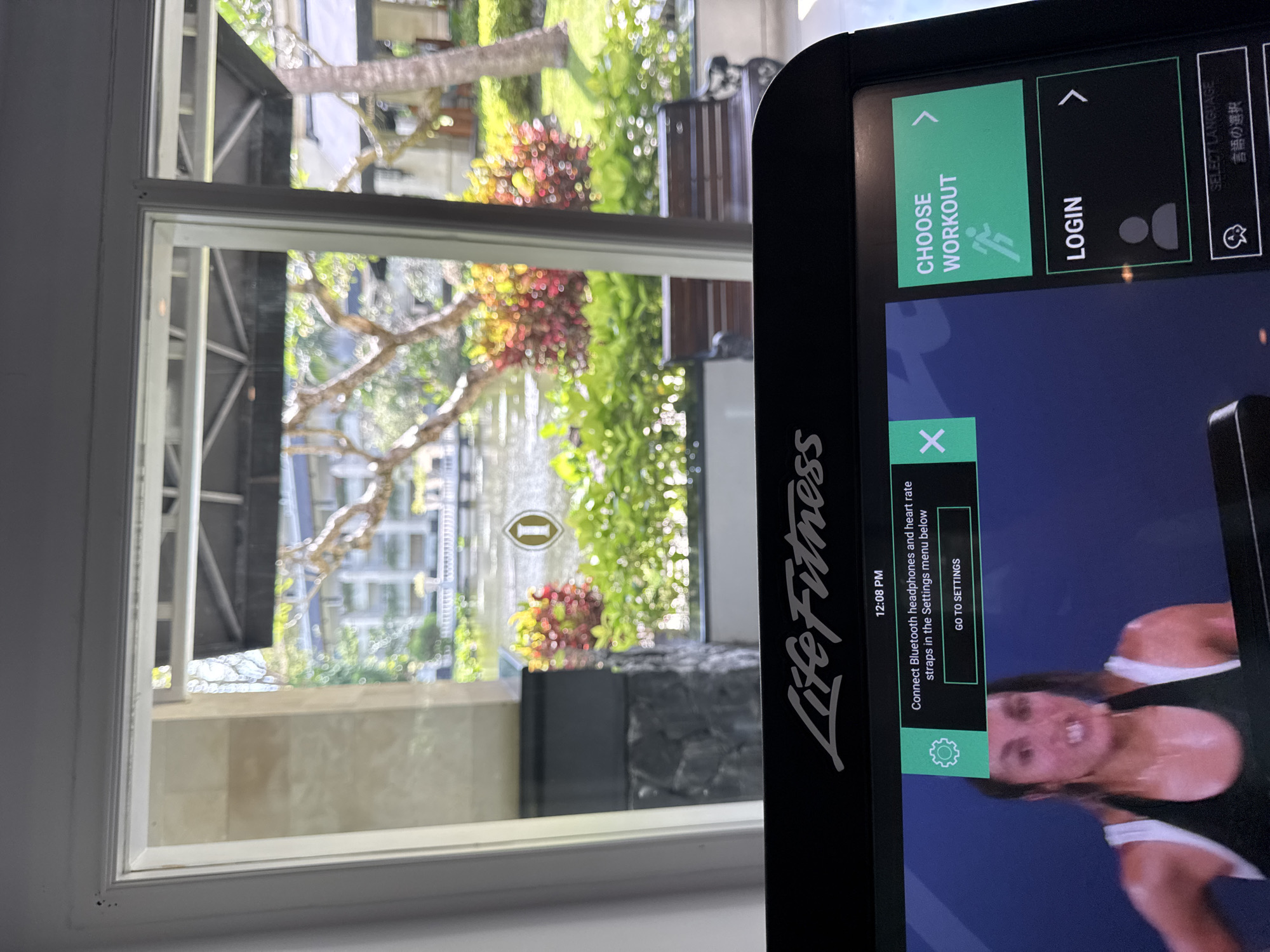 The image shows a view through a window, partially obstructed by the screen of a Life Fitness exercise machine. The window reveals a vibrant outdoor scene with trees, green foliage, and some red flowering plants. In the background, there are buildings that suggest an urban or resort environment. The Life Fitness machine's screen displays options to "Choose Workout" and "Login" and has a notification about connecting Bluetooth headphones and heart rate straps. There is also an image of a woman working out. Based on the context (including the name of the city of Kuta Selatan, Indonesia) the scene suggests a gym or fitness facility located in a tropical environment, likely in a resort or urban area.