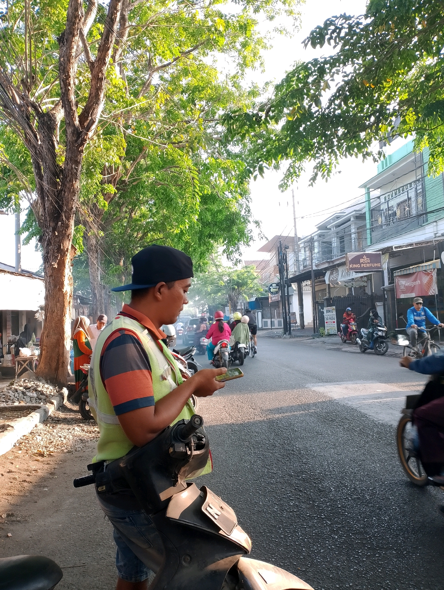 A man wearing a black cap and a reflective vest over a striped shirt stands next to a motorcycle, looking at his phone. He is on the side of a street in Probolinggo, Indonesia. Several other motorcycles and a bicycle are present on the road, with people riding them. Buildings line the street, and trees with green leaves are visible. The scene appears to be outdoors during the daytime, with sunlight casting shadows.