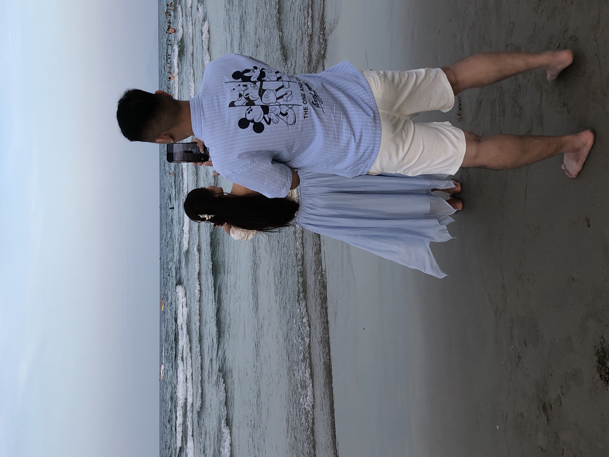 The image depicts a couple on a beach, with the sea and a hazy sky in the background. The couple, positioned on the sandy shore, are facing away from the camera.
The male subject is wearing a light blue and white striped short-sleeved shirt with a graphic print of Mickey Mouse characters and text on the back. He is also wearing light-colored shorts. He is holding a smartphone, appearing to take a picture or video.
The female subject is wearing a light blue dress with a flowing skirt. Her long, dark hair is visible, and she has a white flower accessory in her hair. She is also holding a smartphone, seemingly capturing a moment. Both individuals are barefoot.
The scene is a beach, with waves gently rolling onto the shore. In the distance, there are some boats and people visible in the water, suggesting it's a popular beach. The lighting suggests it might be late afternoon or early evening, with an overcast or slightly hazy sky. The overall mood appears relaxed and casual, with the couple enjoying their time together by the sea. The location is identified as Đà Nẵng, Vietnam.