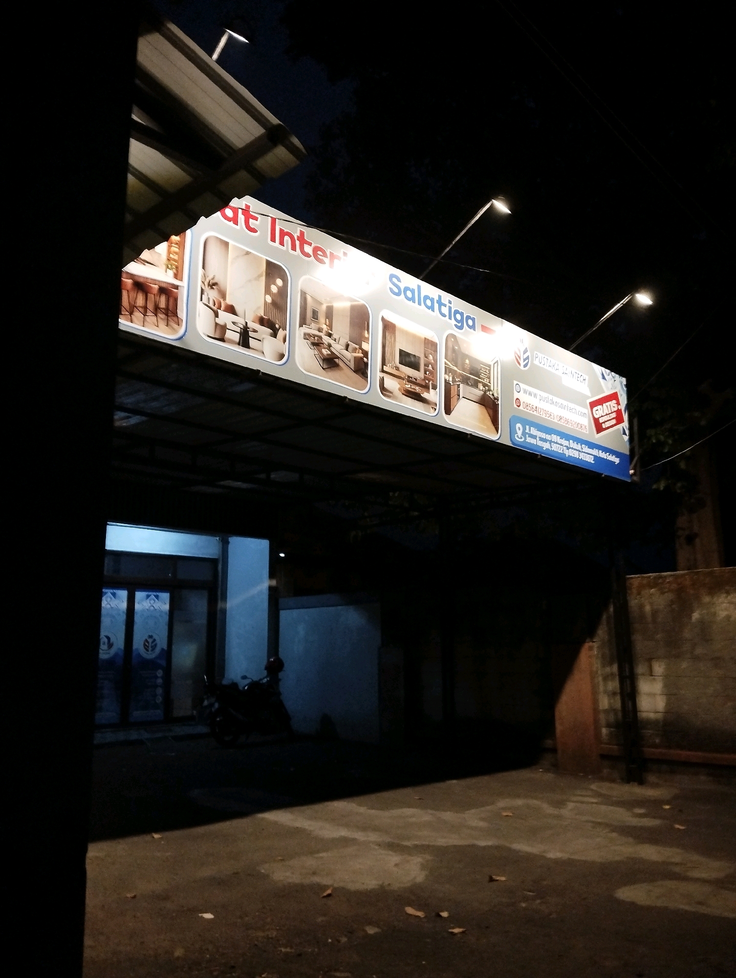 An outdoor, eye-level, nighttime shot shows a brightly lit billboard advertising interior design services in Salatiga, Indonesia. The billboard, which displays images of modern living rooms and kitchen interiors, is mounted on the side of a building and illuminated by overhead streetlights. Text on the billboard includes "Salatiga" and "Pustaka Saintech," along with a website address and phone number. Below the billboard, the entrance to the building is visible, featuring glass doors with logos. A motorcycle is parked in front of the building. The scene is set on an asphalt surface, with some debris scattered on the ground.