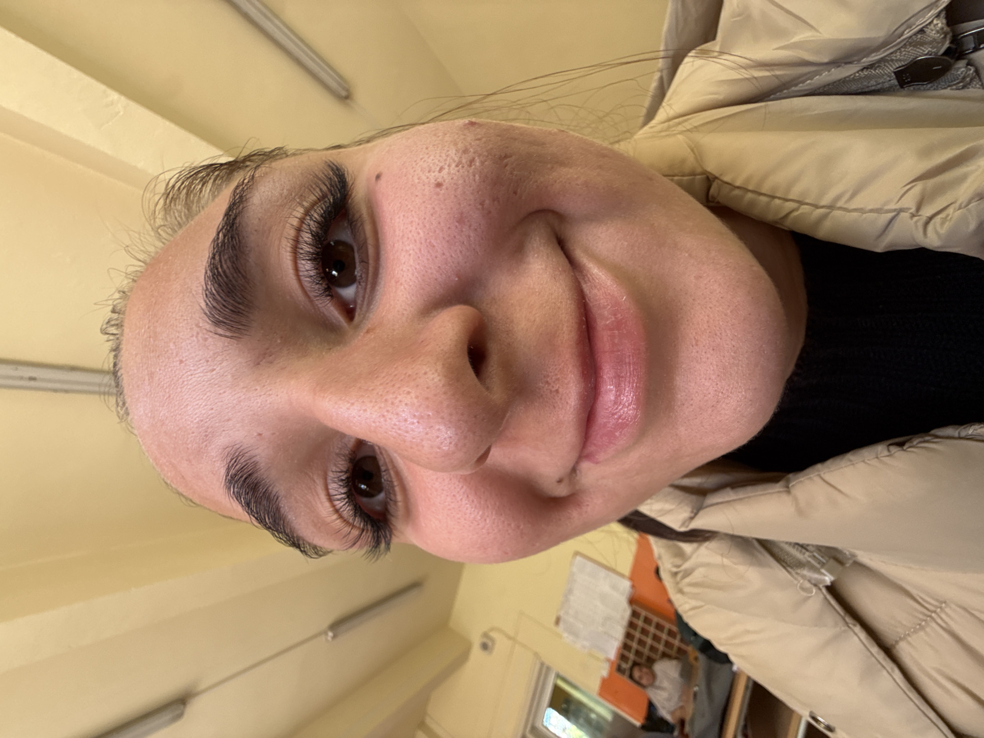 The original image is a close-up portrait of a young person, rotated 90 degrees counter-clockwise. The subject, likely female, is shown from a low angle, looking slightly upwards and smiling warmly. They have dark eyes, full dark eyebrows, and prominent long eyelashes. Their skin displays natural texture, including visible pores and a few small bumps on the forehead and bridge of the nose. A small mole is present above their upper lip. The person is wearing a light beige or tan puffer jacket over a black top, with strands of dark hair visible around their face.
The setting is indoors, appearing to be a classroom or an office in Sofia, Bulgaria. The walls are a light yellow-beige color, and fluorescent strip lighting fixtures are visible on the ceiling. In the background, a window, a partial notice board or papers on a grid, and the indistinct figure of another person are subtly visible. The lighting is artificial and bright, suggesting an indoor environment.
The activity depicted is the subject posing for a photograph, likely a selfie, given the close perspective. Their smile conveys a relaxed and happy emotion. There is no discernible text visible within the frame.
