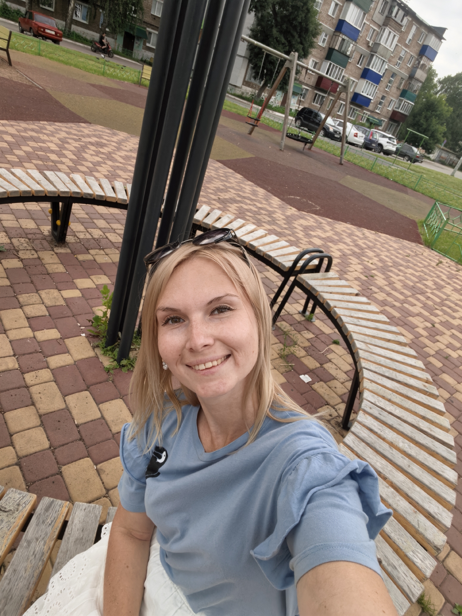 A blonde woman wearing a blue t-shirt and white skirt is sitting on a curved wooden bench in a park. She is smiling at the camera and has sunglasses pushed up on her head. In the background, there are swings and a building. The scene is located in Sterlitamak, Russia.