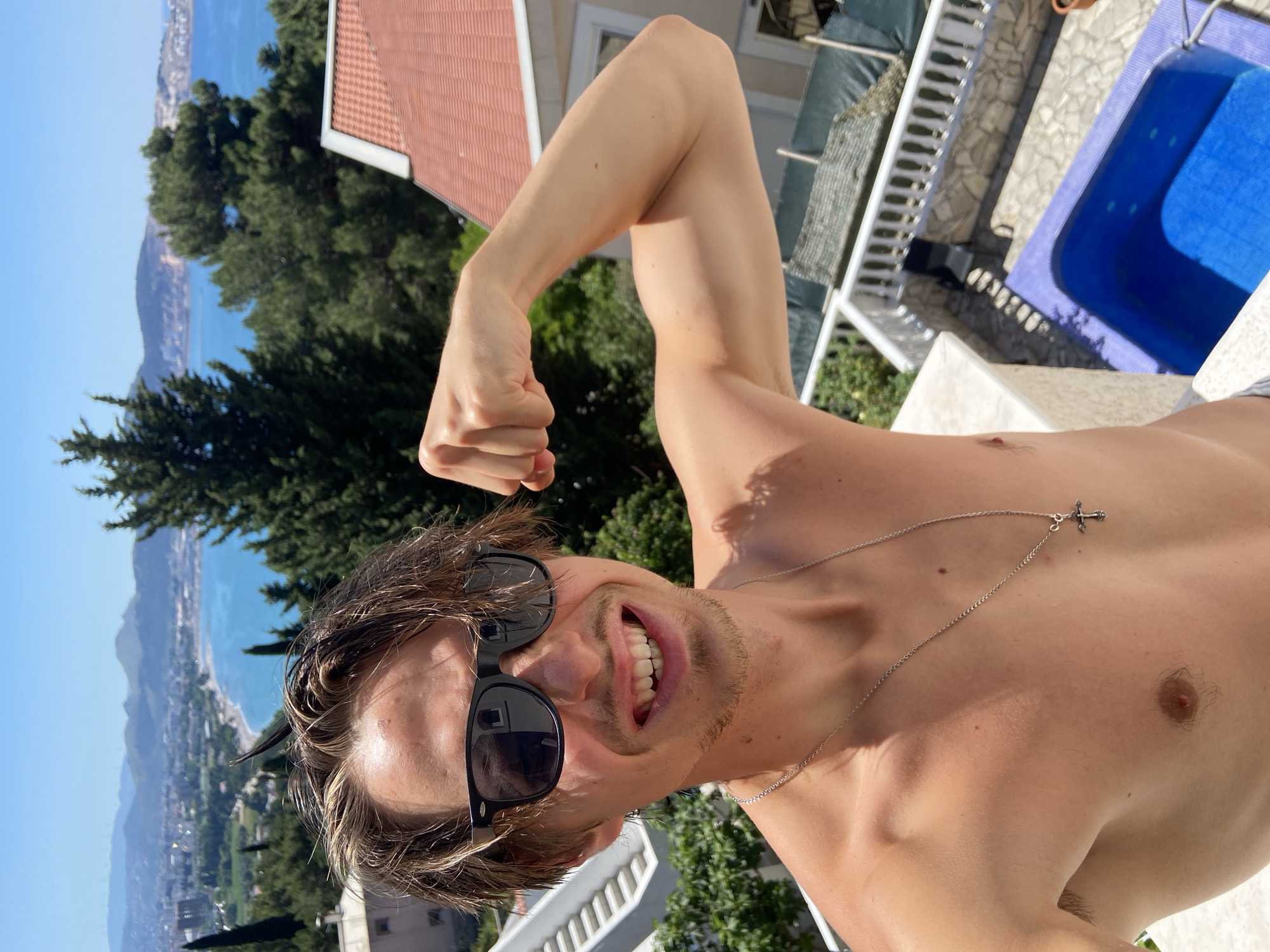 A shirtless man with sunglasses and a necklace poses flexing his arm. He has brown hair and a slight beard. In the background are trees, a building with a red roof, and the sea. A swimming pool is visible to the right of the man. The city in the background appears to be Šušanj, Montenegro, given the coastal terrain and built structures.