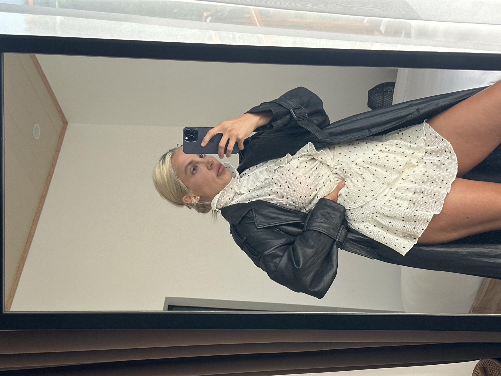 A woman with blonde hair styled in a bun takes a mirror selfie in a room in Kuta Selatan, Indonesia. She is wearing a white, polka-dot dress with ruffled detailing and a black leather trench coat. Her right hand is on her hip, and she is holding a smartphone in her left hand, using it to capture the reflection. The room has light-colored walls, and a large mirror dominates the frame. The background shows a window with sheer curtains and hints of a bed.