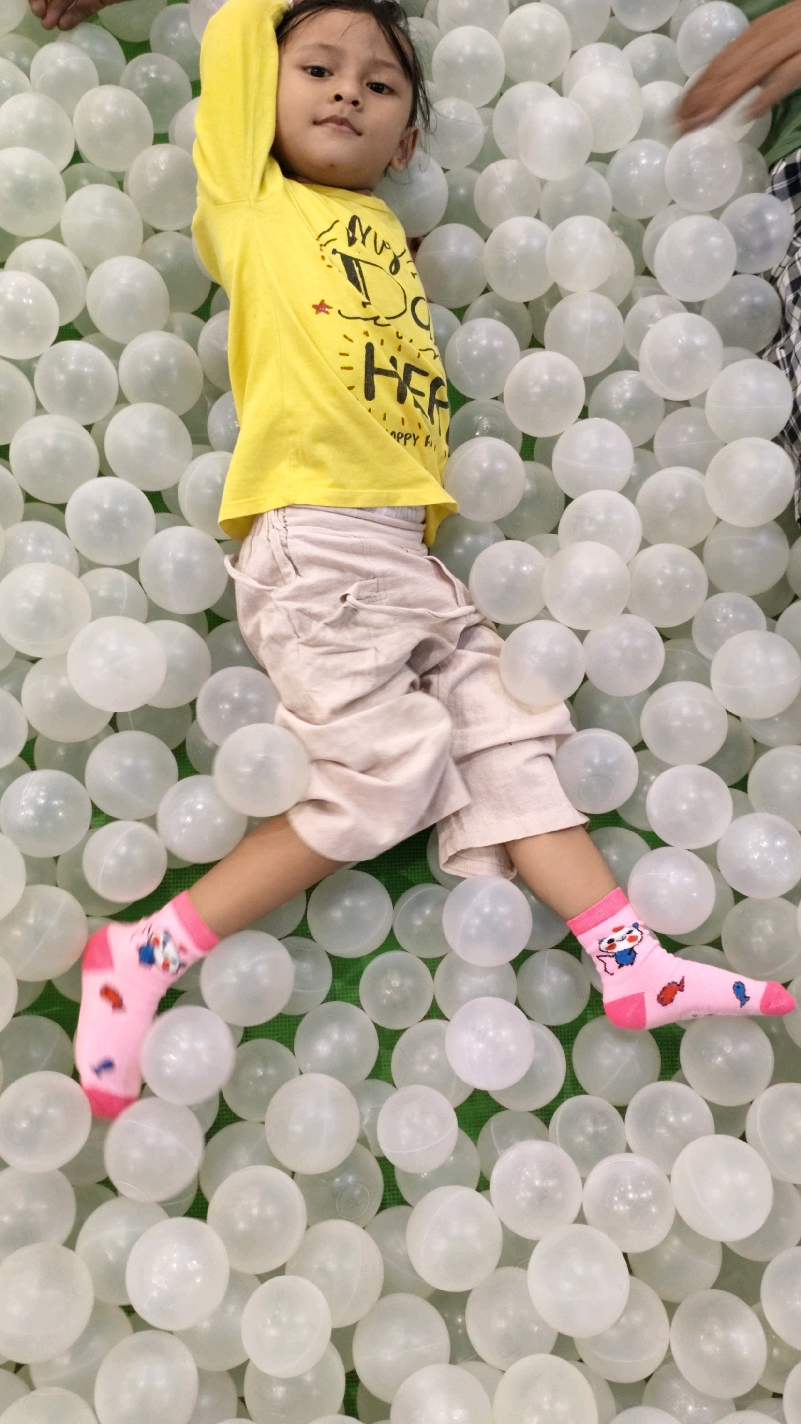 A child in a yellow long-sleeved shirt and light-colored pants is lying on their back in a ball pit. The child is wearing pink socks with cartoon characters. The ball pit is filled with white translucent balls. The background is green. The image appears to be taken indoors, possibly in a play area or a home. The location is not specified in the image or metadata.