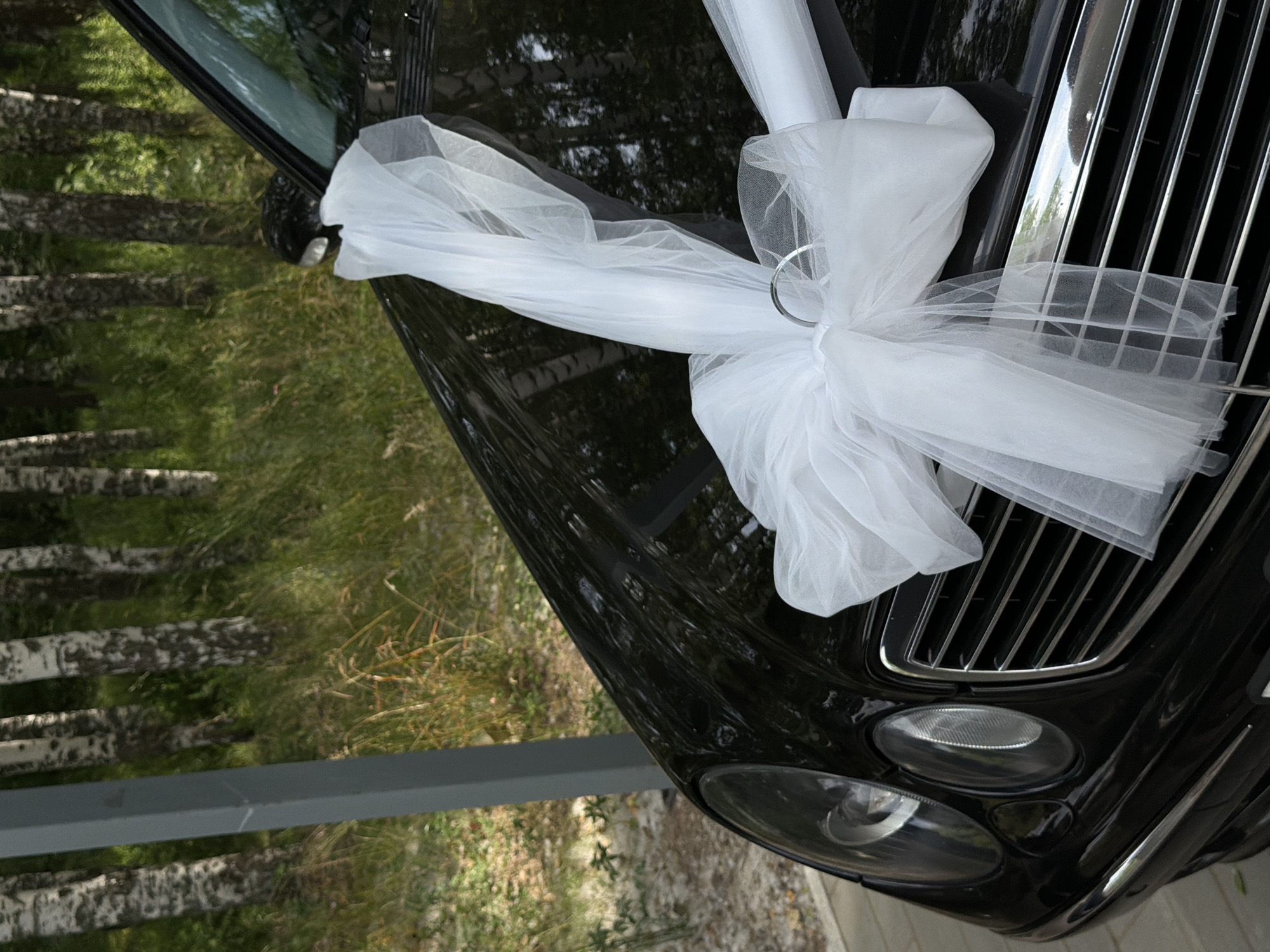 A black Mercedes-Benz E-Class sedan, decorated with a large white tulle bow and ribbon, is parked outdoors in Chaikovsky, Russia. The car is positioned at an angle, with its front left side visible. The bow is centered on the hood, and the ribbon extends towards the windshield. The car's headlights and grille are prominent features. In the background, a row of birch trees and green foliage suggest a natural, possibly wooded, setting. A gray, vertical pole, part of an overhead structure, is visible on the left side of the image. The ground appears to be paved with light-colored material. The overall scene suggests a celebratory or ceremonial occasion, likely a wedding, given the car's adornment.