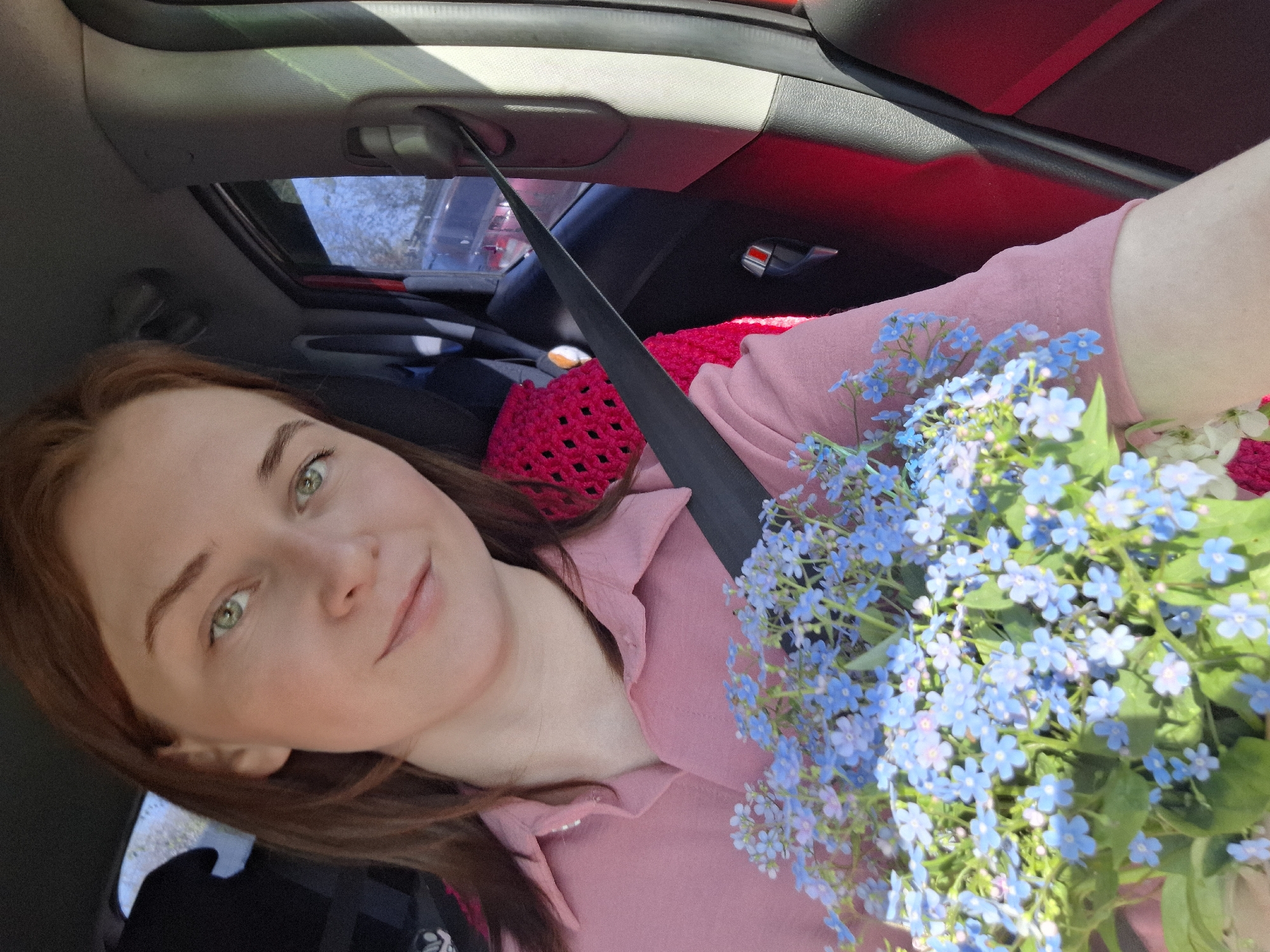 A woman is seated in a car, wearing a pink shirt, with a seatbelt across her chest. She is holding a bouquet of blue flowers, possibly forget-me-nots. The car's interior is visible, with red accents and a sunroof. The scene appears to be inside a vehicle, the location in the city of Chaikovsky, Russia is not possible to determine from the provided image.