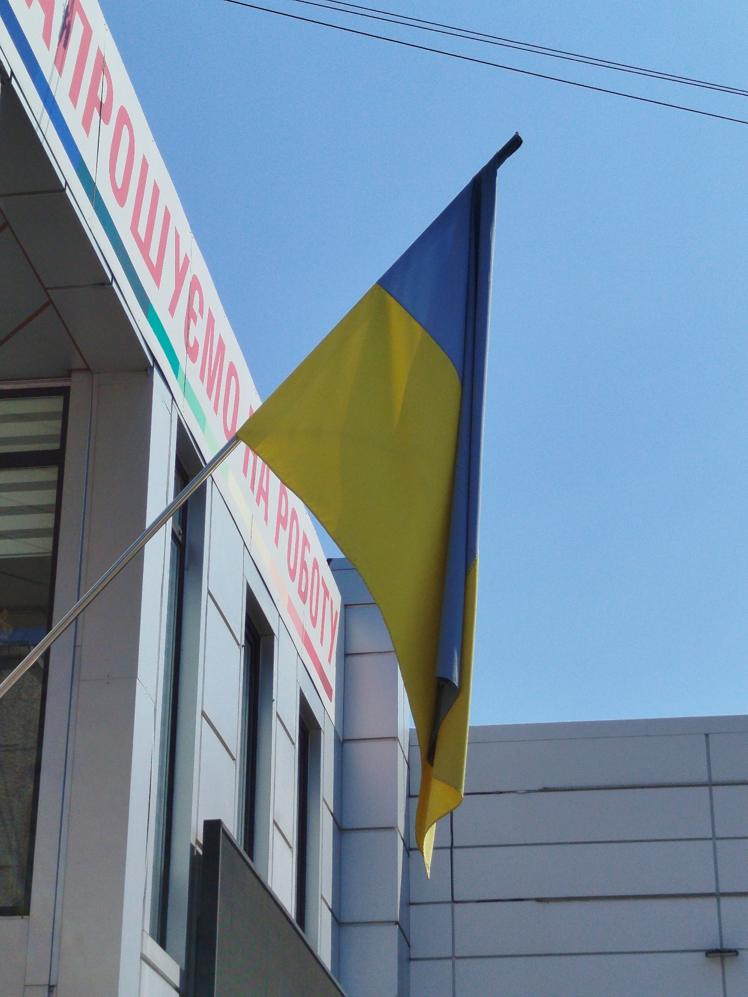 An outdoor, eye-level, medium shot shows the Ukrainian flag against a clear, light blue sky. The flag is positioned on the right side of the frame and is partially obscured by the building on the left. The flag is composed of two horizontal bands, the top band being blue and the bottom band being yellow, with the blue band appearing to be on the hoist side and the yellow on the fly side as the flag blows to the right. The flag is attached to a silver pole that extends from the left side of the frame.
The building on the left is modern, with a light-colored facade and large, dark-framed windows. There is text on the building in red Cyrillic letters, partially visible as "ПРОШУЄМО" and "НА РОБОТУ". The building on the right is also modern with a similar light-colored facade, but it is less detailed in this shot. The overall scene suggests an urban environment. The image was taken in Kremenchuk, Ukraine.