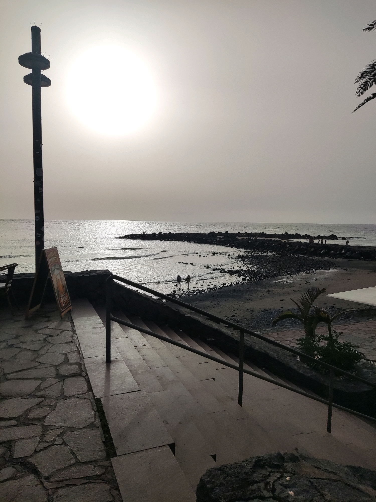 The image captures a view of the beach and ocean at Playa de Las Américas, Spain, under a bright, hazy sun. In the foreground, a stone-paved area leads to a set of concrete stairs with a dark metal handrail. To the left, a dark pole with two circular protrusions stands near a chalkboard sign displaying an advertisement for beer with prices. The beach, composed of dark sand and pebbles, stretches along the waterline, with a breakwater made of large rocks extending into the sea. Several figures are visible on the breakwater and walking near the water's edge. Palm fronds from a nearby tree are visible in the upper right corner. The overall atmosphere is subdued due to the hazy sunlight.