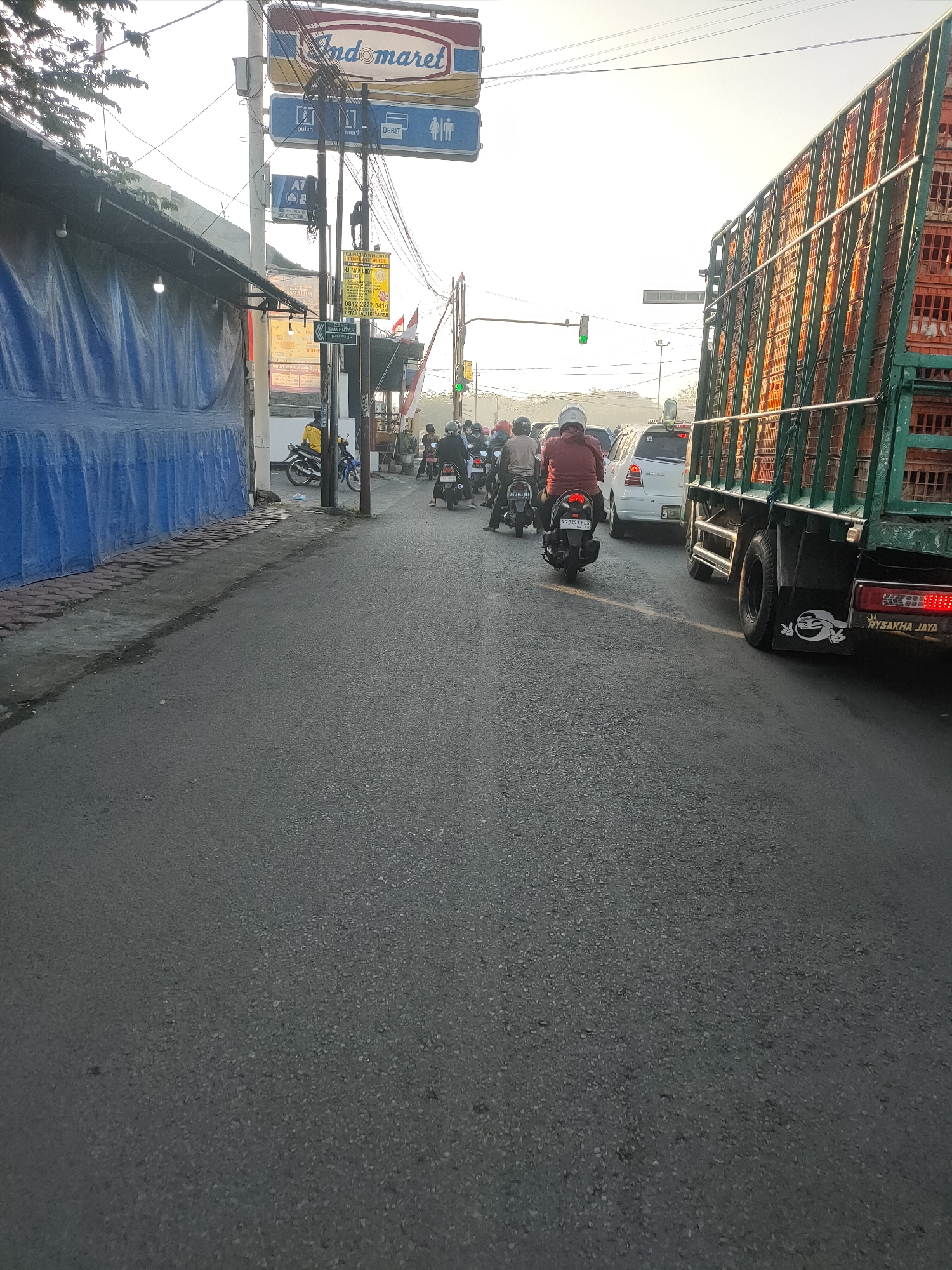 This is a street-level photograph taken in Kanigoro, Indonesia. The scene depicts a congested road with several vehicles, including motorcycles, cars, and a large truck. The truck, on the right side of the image, is carrying a load of orange crates, suggesting it is involved in transportation of goods. Traffic appears to be moving slowly, indicated by the presence of multiple vehicles close together.
On the left side of the road, a building is covered with a large blue tarp. Above the tarp, a sign for "Indomaret," a popular convenience store chain in Indonesia, is visible. This suggests the presence of commercial activity in the area. Other signs, including one for "ATM" and directional signs with icons for restrooms and accessibility, are also present.
Several people are riding motorcycles, some with passengers, in the middle of the road. The lighting suggests it is either early morning or late afternoon, with the sun low in the sky and casting a bright haze. The road surface is asphalt, and the surrounding environment appears to be an urban or semi-urban area. The overall impression is of a busy, functional street in Indonesia.