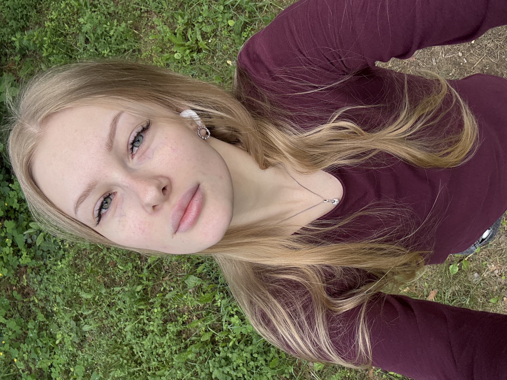 A young woman with long blonde hair and light blue eyes is captured in a high-angle, selfie-style shot outdoors. She is wearing a burgundy long-sleeved shirt and a silver necklace with a clear pendant. She also has a small, circular earring with small stones in her left ear and a white wireless earbud in her right ear. The background is a dense, green grassy area with some patches of dry leaves and dirt, suggesting a natural, outdoor environment possibly in a park or wooded area within the city of Perm, Russia. Her facial expression is neutral, and she is looking directly at the camera. The lighting is natural daylight, with no artificial light sources visible.