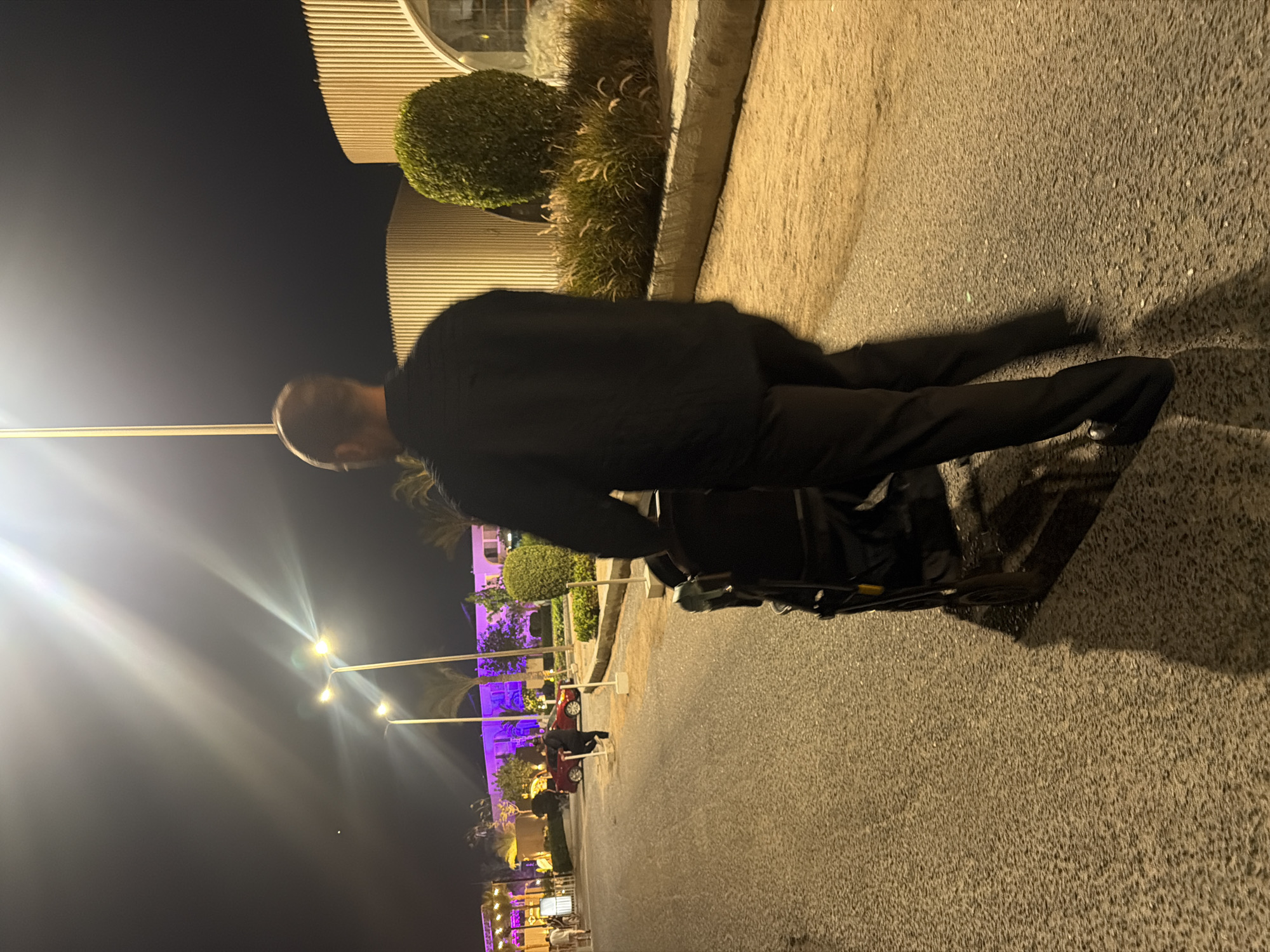 A man wearing a black shirt and pants pushes a stroller down a paved street at night. The street is lined with trees and modern buildings with white vertical siding. Streetlights illuminate the scene with a bright, warm glow, casting long shadows. In the background, there are more lights, suggesting a lively area, possibly a resort or entertainment district in Sharm El Sheikh, Egypt.