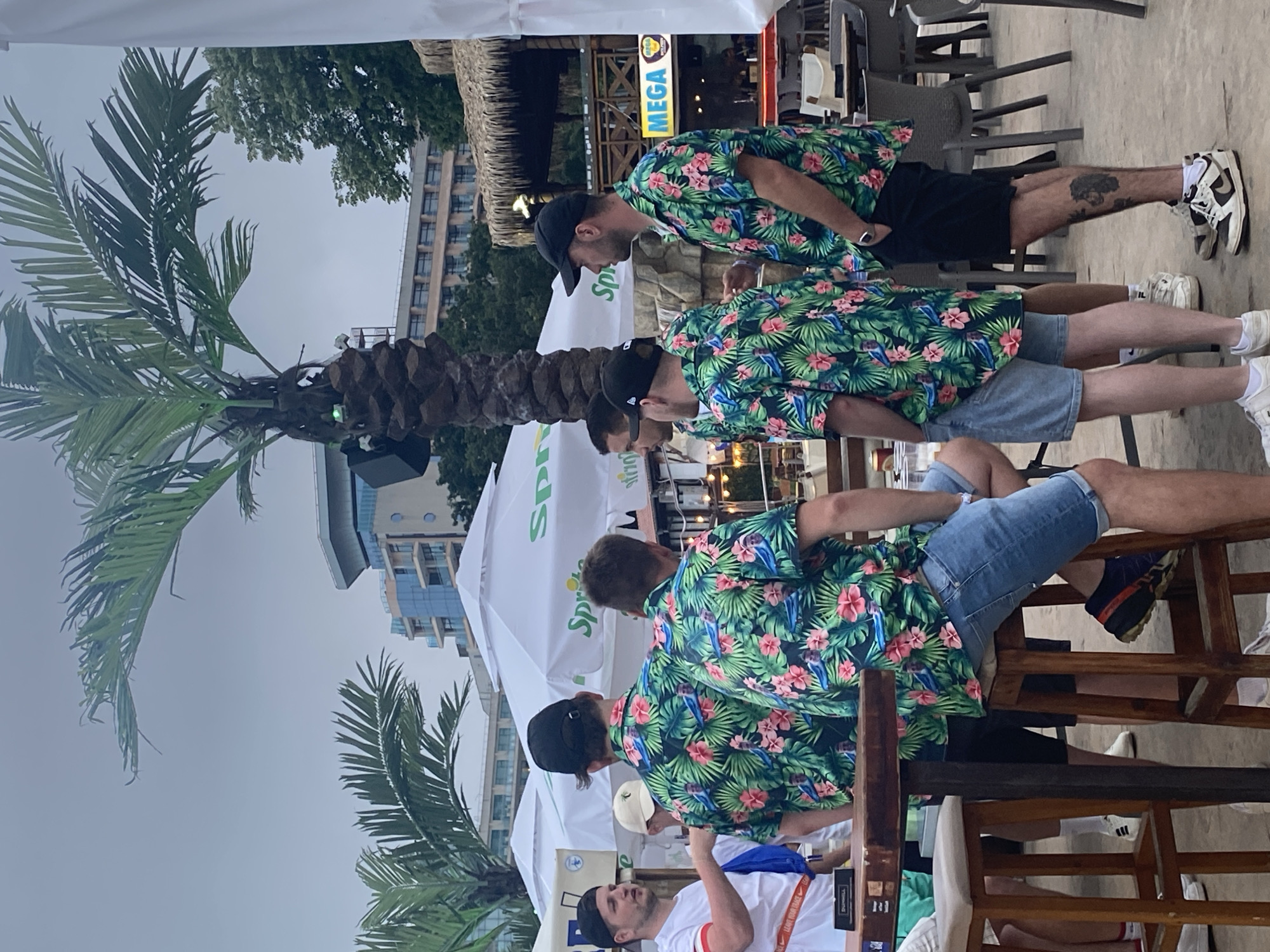 An outdoor scene in Varna, Bulgaria, depicts a casual gathering of several individuals under an overcast sky. The foreground is dominated by a group of at least seven men congregated around a wooden bar-height table with matching stools. Five of these men are prominently featured. Four of them wear identical short-sleeved green shirts with a tropical floral pattern featuring pink flowers, green leaves, and blue birds.
From left to right:
1. A white male, wearing a white short-sleeved t-shirt with red trim, a black backwards baseball cap, and light-colored shorts. He stands, facing right, with his left arm extended towards the table. Text "NIKE 'LEAVE YOUR TRACE'" is visible on his shirt.
2. A white male, seen from the back, wearing a black backwards baseball cap and one of the tropical shirts. He stands behind the wooden table.
3. A white male, seated on a wooden stool with his back mostly to the camera, wearing a light-colored baseball cap and one of the tropical shirts. He is wearing light-wash denim shorts.
4. A white male, seated on a wooden stool, seen from the right side, wearing a black baseball cap and one of the tropical shirts. He is wearing light-wash denim shorts and dark blue athletic shoes with an orange sole.
5. A white male, standing, seen from the right side, wearing a black baseball cap and one of the tropical shirts. He is wearing dark shorts, white socks, and black and white sneakers, with a dark tattoo visible on his right calf.
In the background, multiple large palm trees are visible, one of which has visible speakers or lights attached to its trunk. Several white outdoor umbrellas, branded with a green "Sprite" logo and text, provide shade. Beyond these, modern multi-story buildings with balconies and glass facades are visible, characteristic of a coastal resort town. To the far right, a structure with a thatched roof is partially visible, featuring a yellow sign with red text "MEGA." The ground surface appears to be paved or a similar outdoor material. The sky is uniformly light gray, indicating an overcast condition or late afternoon/early evening light.