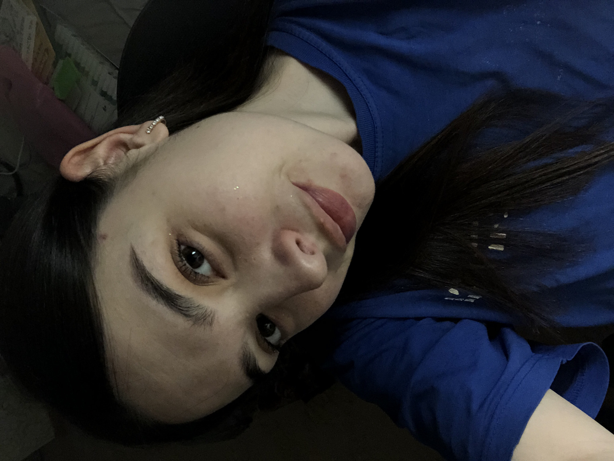 A young woman with dark brown hair is positioned in the lower center of the frame, angled to the left. Her face is partially illuminated, highlighting her features: dark eyebrows, brown eyes, nose, and lips. She is wearing a blue long-sleeve shirt, with the sleeve visible in the lower right corner. A piece of jewelry is visible in her ear. The background is dark and indistinct, with a glimpse of objects in the upper left corner. The location in the city of Noviy, Russia is unknown.