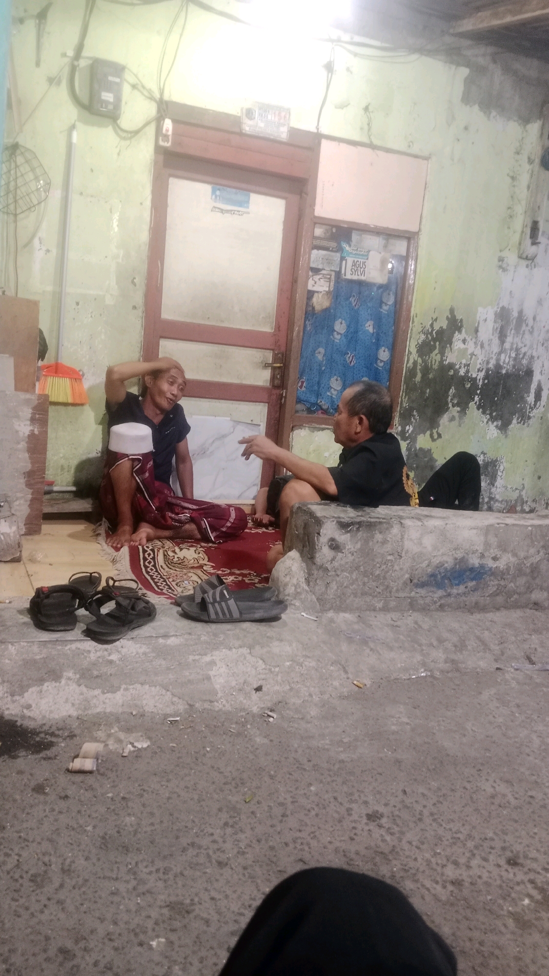 Two men are seen sitting and interacting in what appears to be a courtyard or outdoor common area of a residential building in West Jakarta, Indonesia. The man on the left, wearing a black t-shirt and a white skullcap, is gesturing with his right hand while seated on a red and patterned rug. He is also wearing a maroon patterned sarong. The man on the right, in a black t-shirt and black pants, is reclining on a concrete structure, facing the other man and appearing to engage in conversation. The environment shows signs of age and wear, with peeling paint on the walls and a wooden door visible in the background. Several pairs of sandals are scattered on the ground in the foreground. There are also various debris on the concrete floor, including what appear to be cigarette butts. The overall lighting suggests it might be evening or dimly lit conditions.