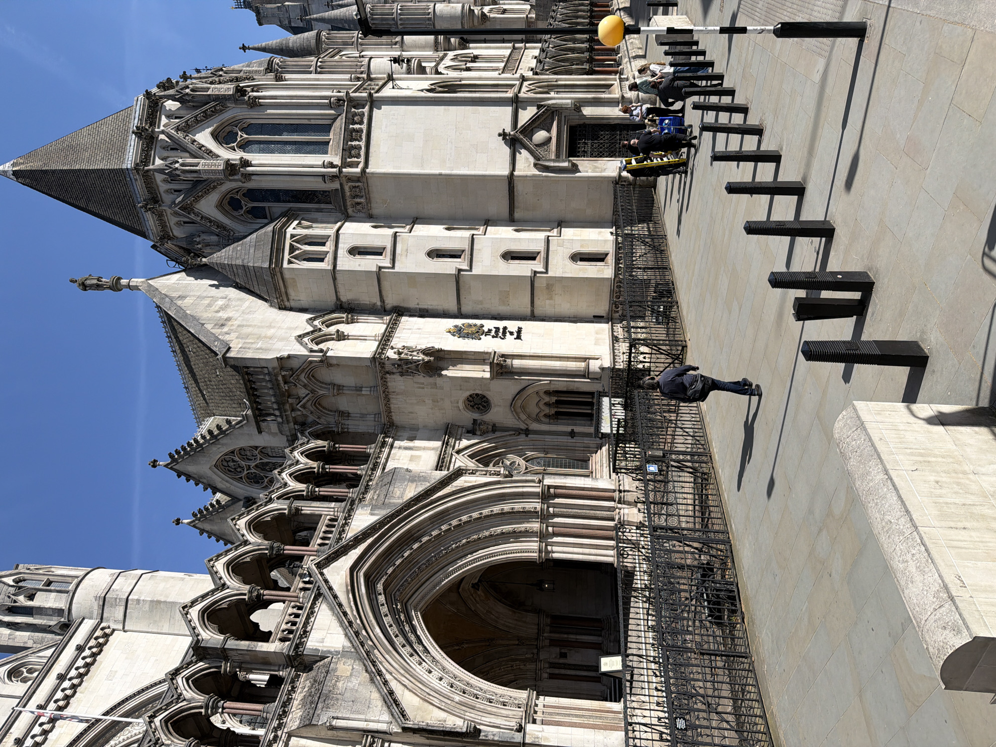 The image shows the Royal Courts of Justice building in London, United Kingdom. The building has a Gothic Revival style with multiple arches, windows, and spires. The facade is made of light-colored stone. A black wrought iron fence runs along the building. On the sidewalk, there are metal bollards with black and white poles. People are sitting on the sidewalk near the building. One person is walking away from the building. The sky is blue with no clouds.