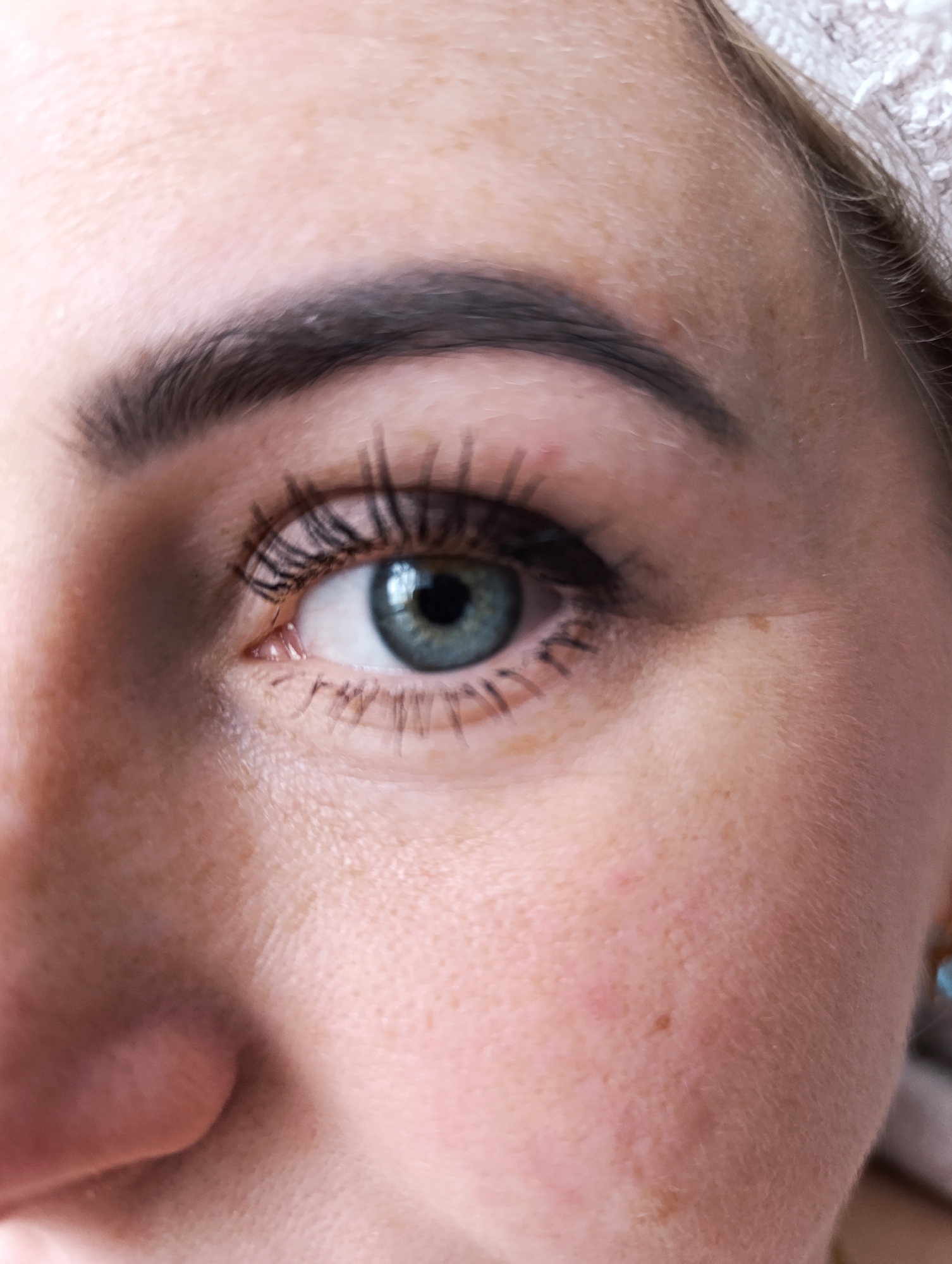 A close-up of a person's right eye, eyebrow, nose, and cheek. The eye is a shade of blue-green, with black mascara visible on the eyelashes. The eyebrow is thick and dark. The skin shows freckles and some redness. The location and city are not discernible from the image.