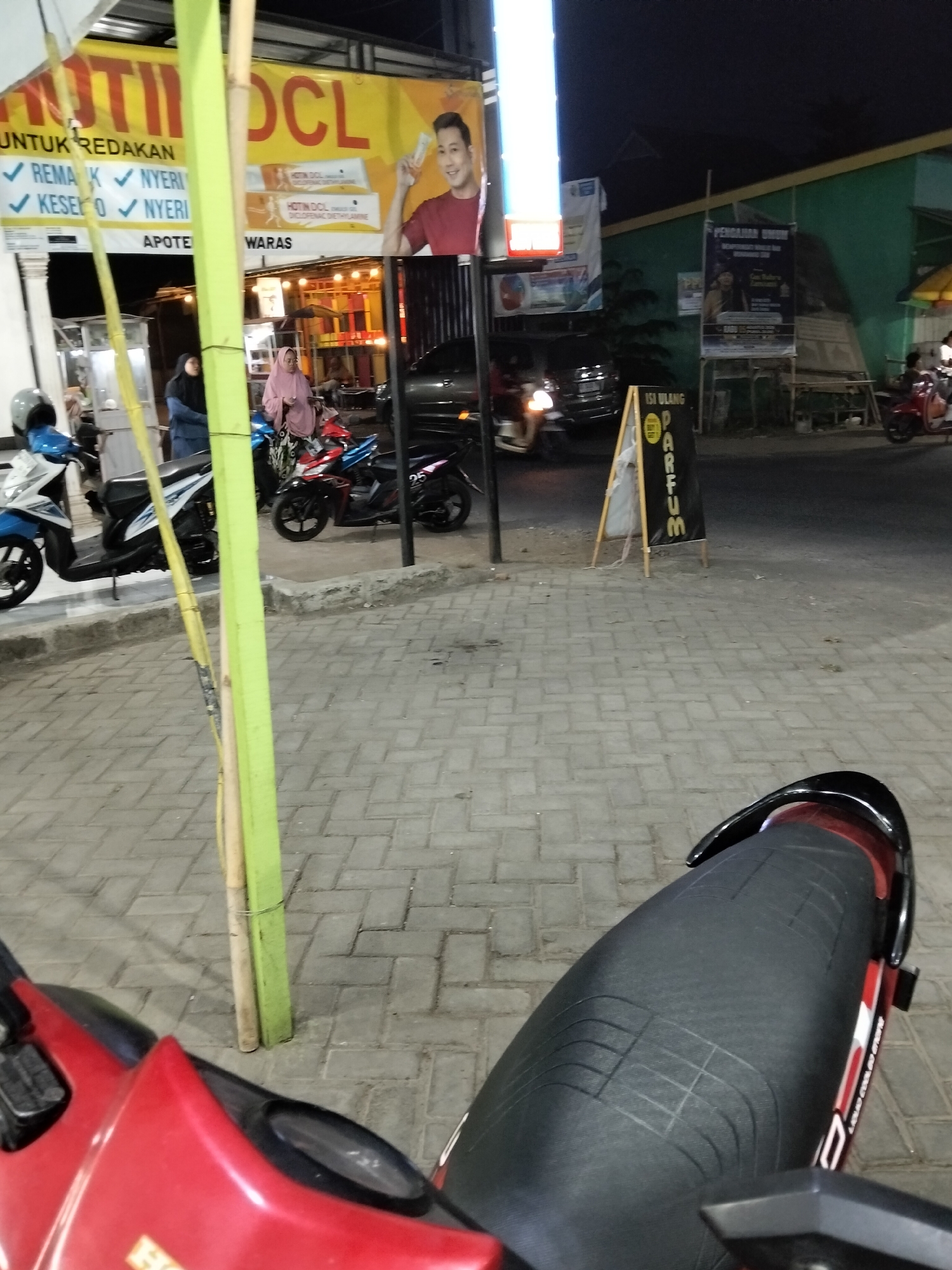 The image shows a street scene at night in Gempolan, Indonesia. In the foreground, a red and black motorcycle is parked. Beyond the motorcycle, the street is paved with interlocking bricks. A bright green and yellow bamboo pole stands vertically in the middle of the image, with a yellow wire wrapped around it.
In the midground, several motorcycles are parked. A blue and white scooter is on the left, and a red and black motorcycle is to its right. Two women, one wearing a pink hijab and the other a dark blue hijab, are standing near the motorcycles. Behind them, there is a building with a large yellow banner that reads "HOTIN DCL" and features an image of a man holding a product. There is also a sign that says "APOTEK WARAS". Further back, a black car is parked.
In the background, there is a green building and a dark sky. Another sign, an A-frame stand, displays "ISI ULANG PARFUM" (refill perfume). Various other signs and structures are visible, indicating a commercial area. The overall impression is of a busy street scene at dusk or night, likely a commercial district in Gempolan.