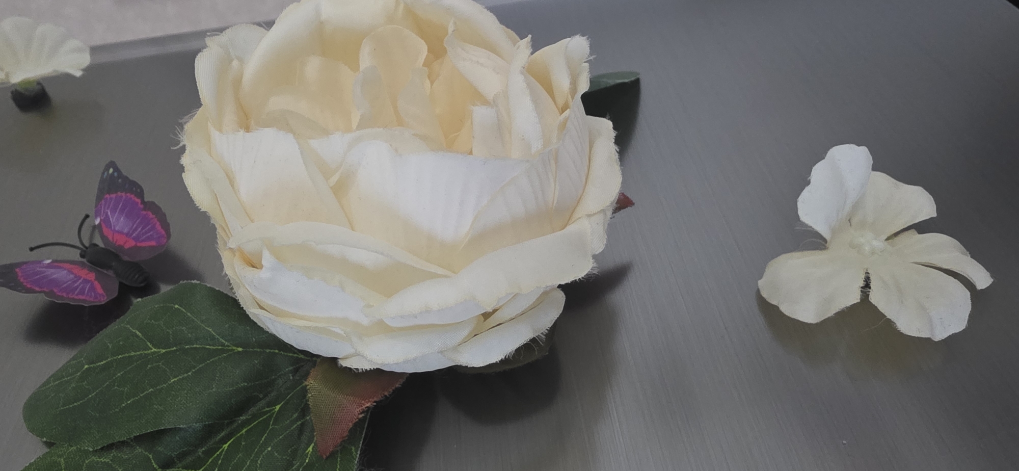 The image shows a close-up of a large, creamy white artificial peony flower and a small, white artificial flower, positioned on a grey, textured surface. A small, dark-bodied butterfly with purple and pink wings is attached to the left side of the peony, near its green leaves. The lighting suggests an indoor setting, with shadows cast by the flowers and butterfly. There are no people visible in the image, nor any indication of the city of Sumgayit, Azerbaijan.
