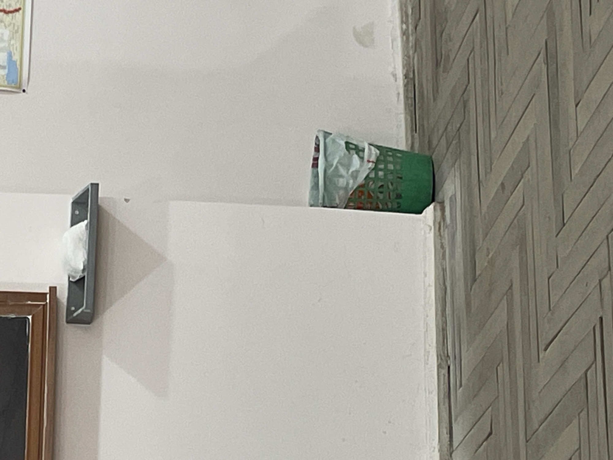 The image shows a section of an interior wall. The left portion of the wall is smooth and painted white. Towards the right, the wall transitions to a decorative pattern featuring grey, interlocking geometric shapes. A small, rectangular green plastic basket is perched on a ledge where the two wall sections meet. Inside the basket, there appears to be some material wrapped in light-colored plastic. To the left of the basket, a tissue holder and a portion of a framed picture are visible. The image does not provide enough information to determine the specific location in Yerevan, Armenia. There are no people visible in the image.