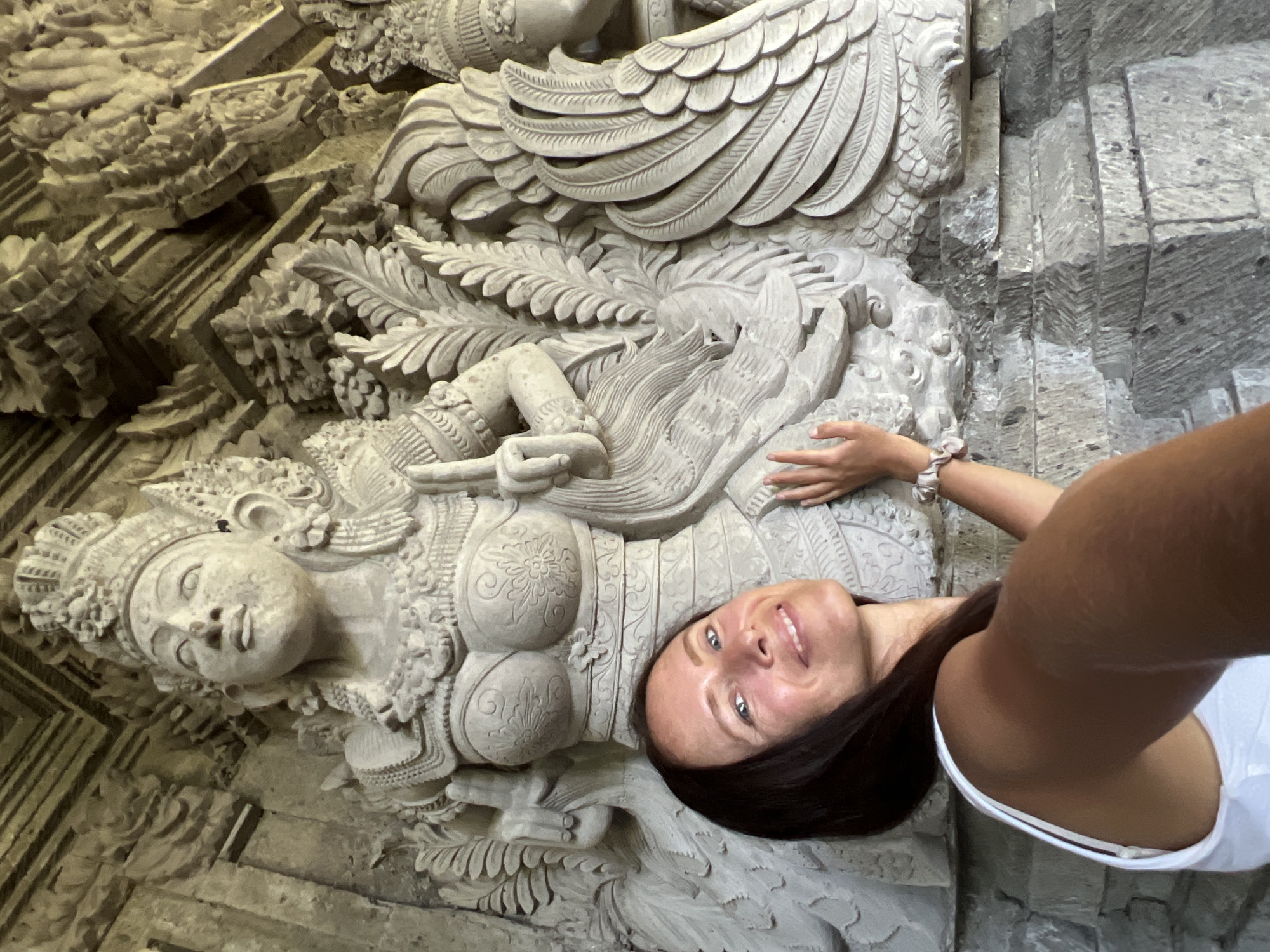 A woman takes a selfie with a bas-relief sculpture in Kedewatan, Indonesia. The sculpture features a female figure with ornate details, including elaborate headdress, chest ornamentation, and flowing garments, and she reclines amidst foliage and winged figures. The surrounding architecture consists of carved stone elements. The woman, with dark hair and a white tank top, smiles at the camera.