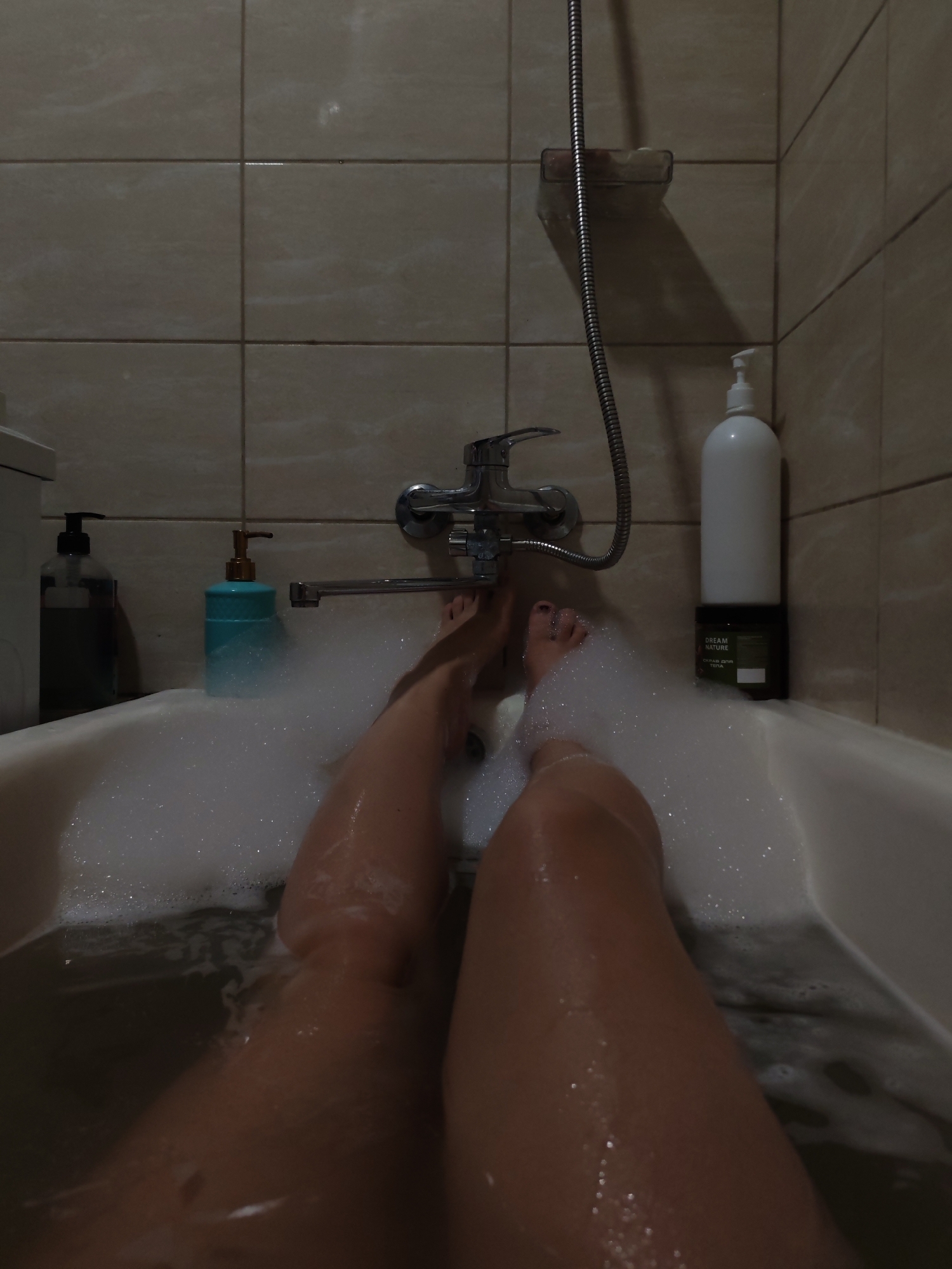 This is a first-person perspective, eye-level shot of a person's legs submerged in a bubble-filled bathtub. The scene takes place in a bathroom with beige tiled walls. On the edge of the bathtub, to the left, are two pump bottles, one clear with dark liquid and the other a teal color. To the right, there is a tall white pump bottle next to a dark green container. A faucet and showerhead are visible on the wall above the tub. The lighting is dim, creating a relaxed and intimate atmosphere. The person is lying down, with their feet slightly visible above the water and bubbles. The specific location in Izhevsk, Russia is not determinable from the visual information alone.