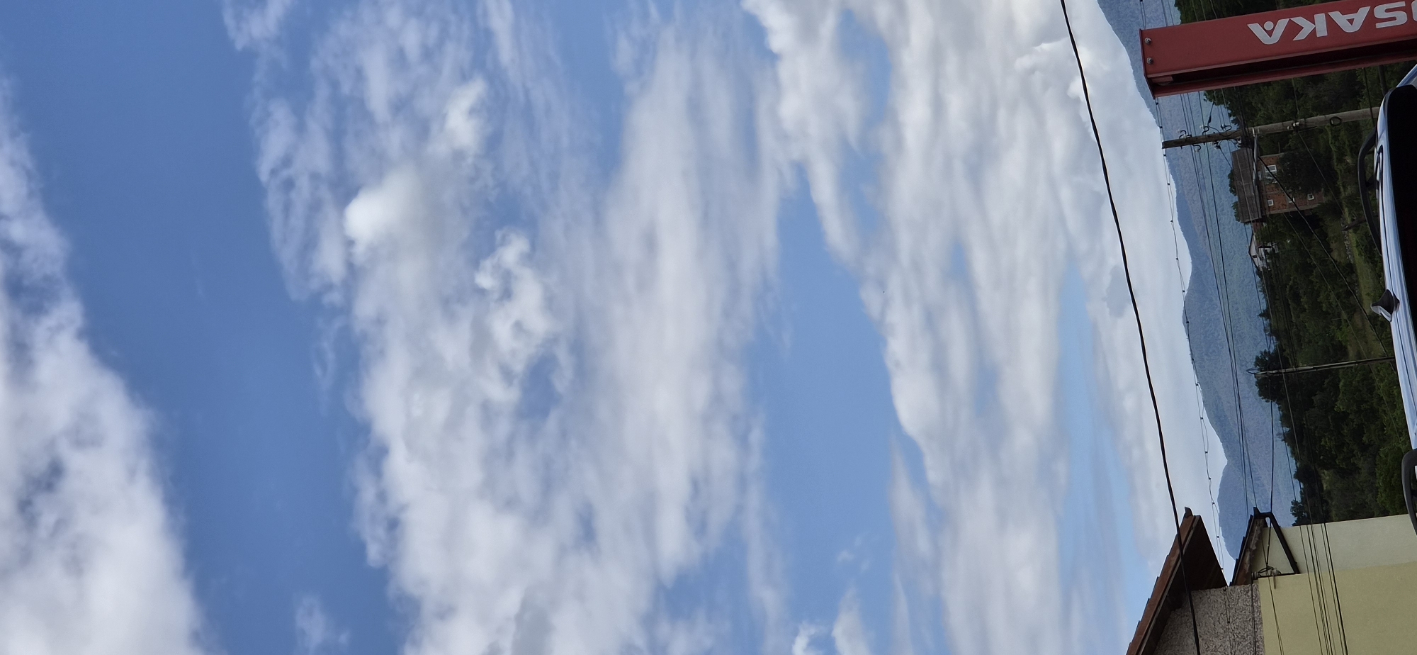 The image depicts a daytime outdoor scene with a bright blue sky partially obscured by white, fluffy clouds. In the foreground, a portion of a building with a light yellow facade and a brown roof is visible on the left. A dark horizontal line, possibly a wire or power line, stretches across the upper mid-section of the image. Below the sky and clouds, a range of green, rolling hills forms the horizon, with some trees and a few buildings scattered amongst them. A tall, red vertical sign with white text that appears to read "OSAKA" is positioned on the right side of the frame, partially obscuring the landscape behind it. A dark-colored vehicle's roof is visible in the bottom center of the image. The overall impression is a rural or suburban setting under a partly cloudy sky. This scene is located in Radilovo, Bulgaria.