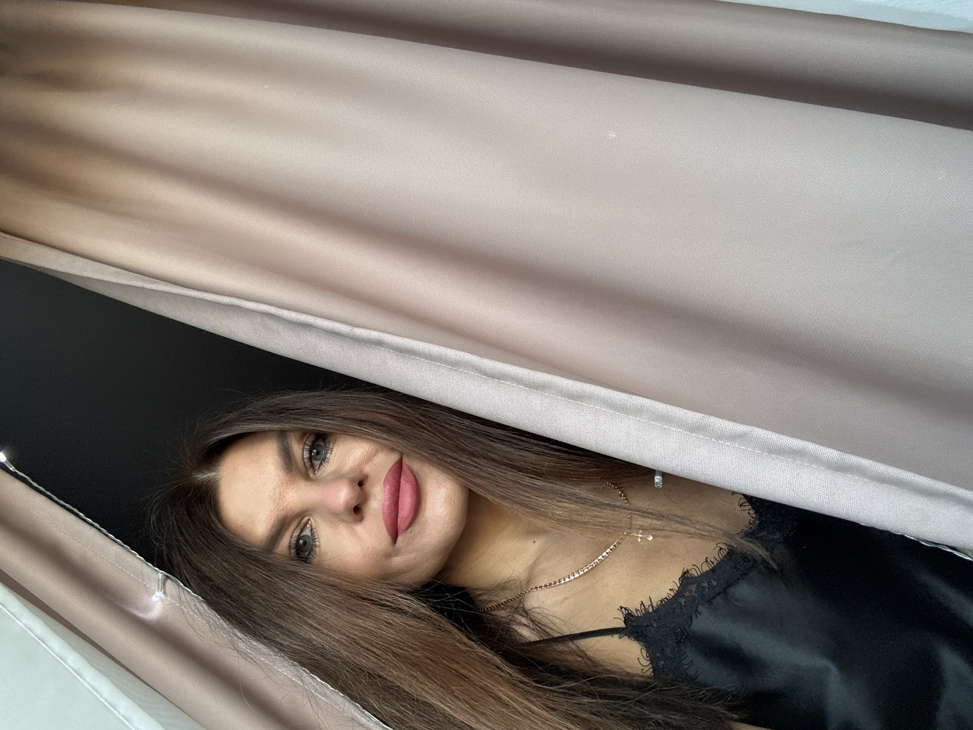 A woman is positioned in front of beige curtains, partially obscuring her. She has long, brown hair and is wearing a black camisole top with lace trim. A thin gold necklace with a cross pendant is visible around her neck. The background behind the curtain appears to be a dark wall. Based on the location information, the image was taken in the city of Chaikovsky, Russia.