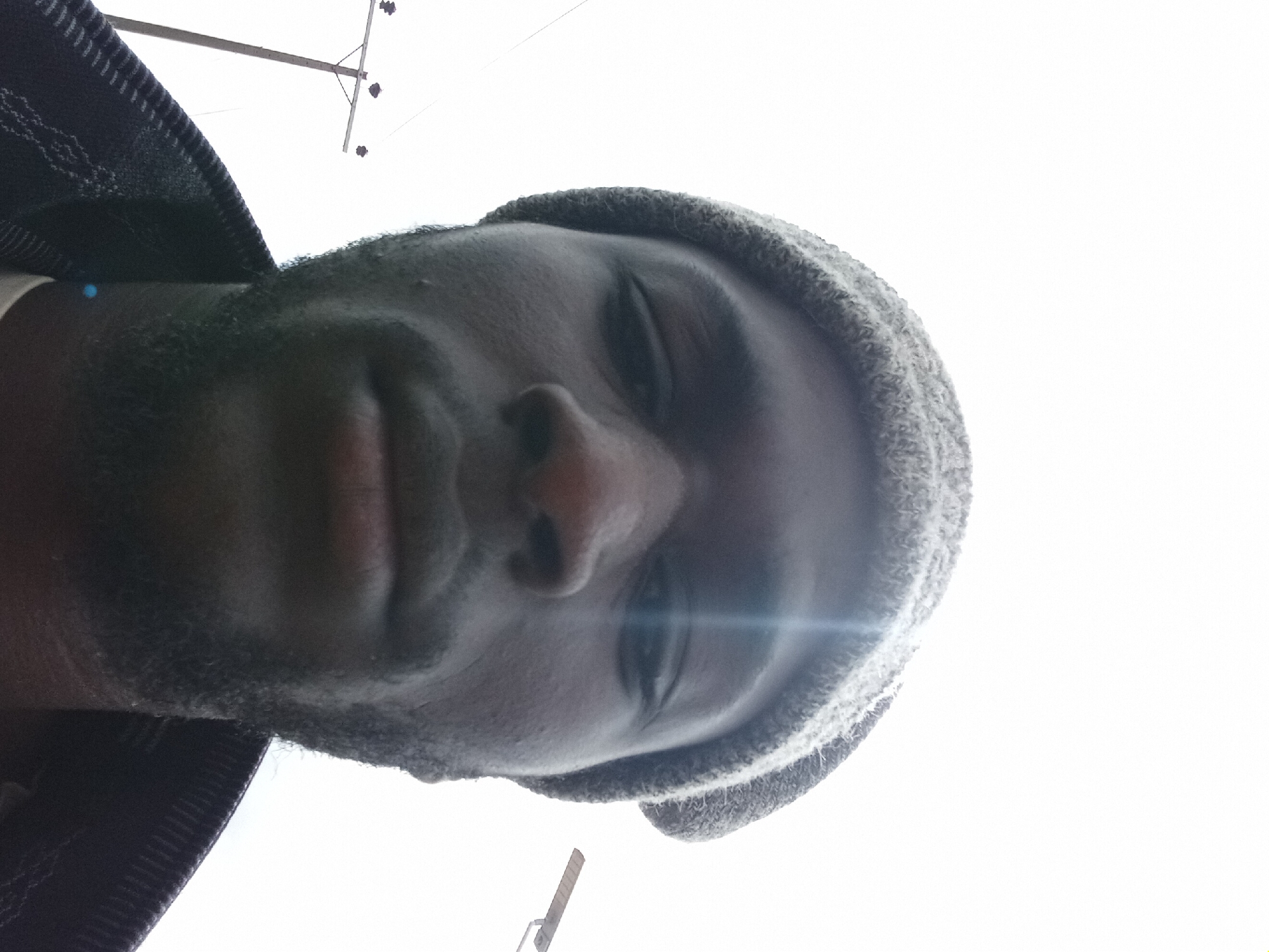The image is a close-up, low-angle selfie of a Black man wearing a gray beanie and a dark jacket. He has a beard and is looking directly at the camera. The background is a bright, overcast sky with a utility pole and street light visible on the right side. The man is positioned in the foreground, with his face filling most of the frame. The lighting suggests it is daytime, and the presence of the utility pole and street light indicates an outdoor urban or semi-urban setting. The location is specified as Bukuru, Nigeria.