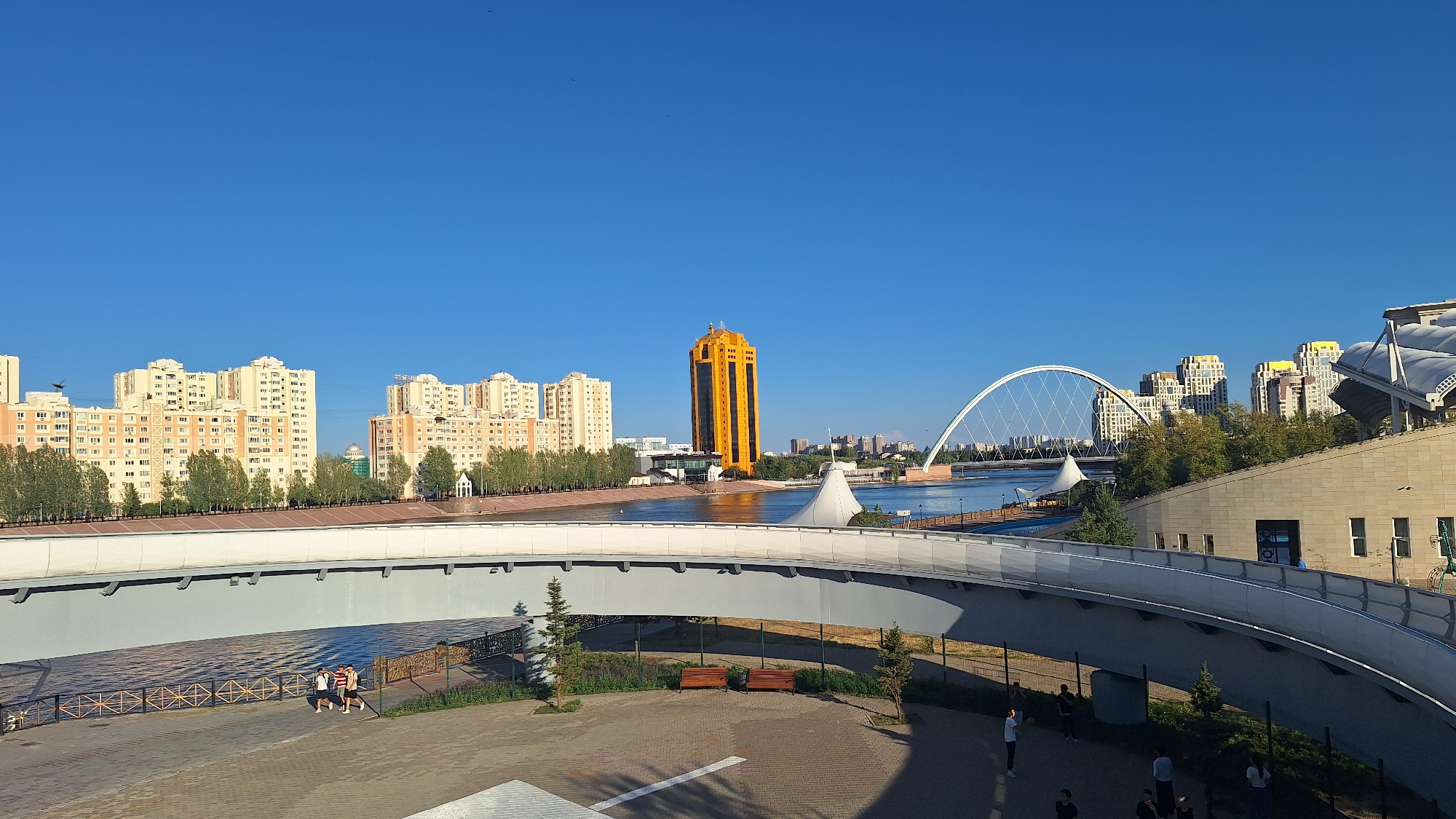The image captures a panoramic view of Astana, Kazakhstan, under a clear blue sky. In the foreground, a modern, grey pedestrian bridge curves across the frame, casting shadows on the paved area below. Several people are visible on the walkway beneath the bridge, including a group of three individuals in athletic wear walking together and another person standing. Two wooden benches are placed on a patch of grass next to the walkway.
Beyond the bridge, the Ishim River flows, reflecting the bright sunlight. On the far bank, a striking orange skyscraper stands tall, its facade shimmering. To the left of the skyscraper are several light-colored apartment buildings with numerous windows. Further to the right, a distinctive white arched bridge spans the river. Behind the arched bridge, more apartment buildings with yellow accents on their rooftops rise against the skyline.
To the right of the arched bridge, a modern building with a beige stone facade and a white, tent-like roof structure is visible. Trees line the riverbanks, adding greenery to the urban landscape. The scene suggests a pleasant day in the city, with people enjoying the outdoor spaces. The architectural styles and the presence of the Ishim River, the arched bridge (likely the Water-Land Bridge), and the orange skyscraper (possibly the KBTU building) strongly indicate the location as Astana, Kazakhstan.