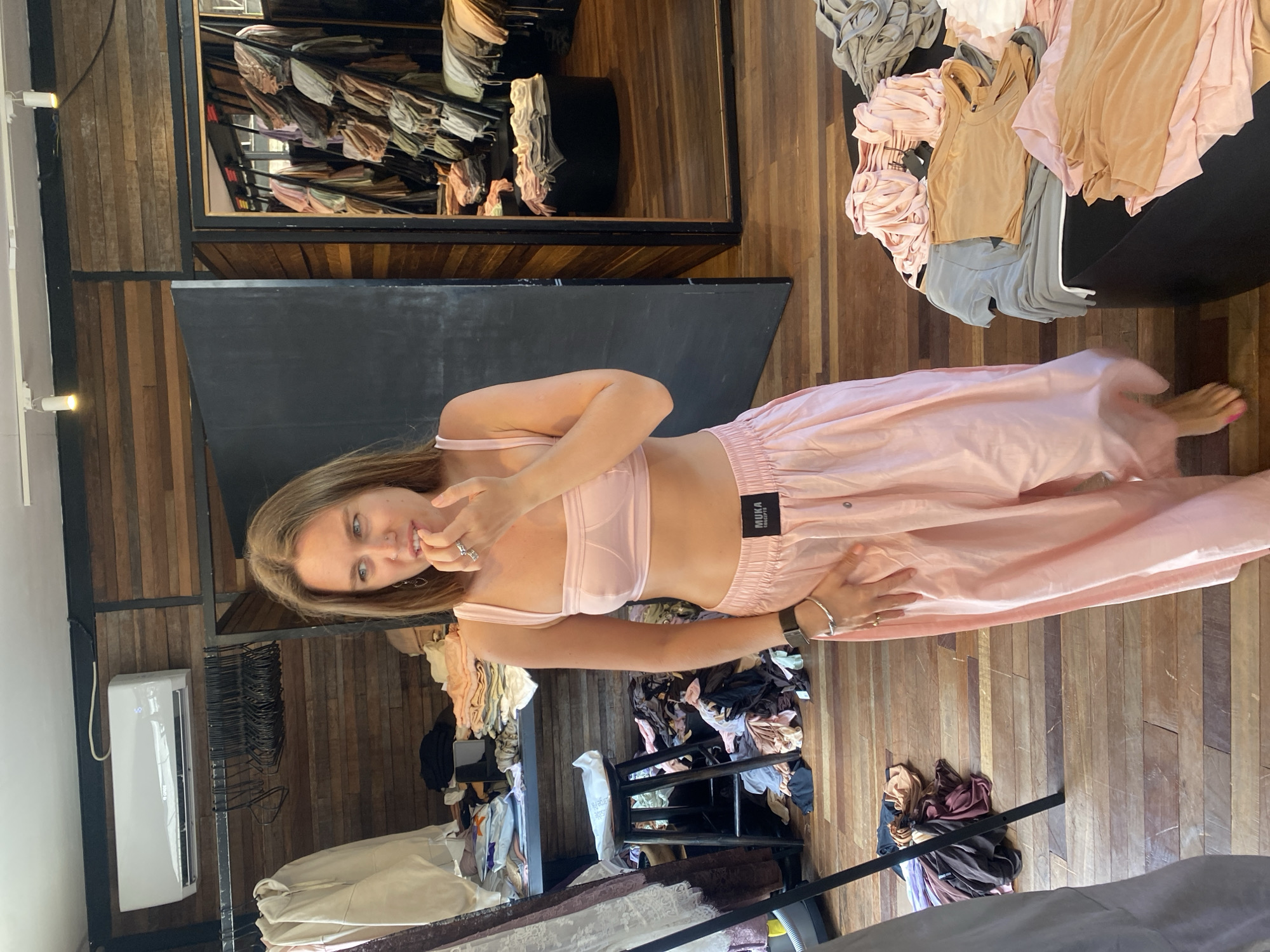 A young Caucasian woman with long, light brown hair stands in a cluttered clothing store. She is wearing a light pink bra top and loose-fitting pink pants. Her right hand is touching her mouth, and she is looking to her left. The store has wooden walls and floors, with shelves of clothing visible in the background. There is also a large mirror and an air conditioning unit mounted on the wall. The area appears to be a retail space filled with clothing racks and folded garments. The woman's attire suggests she might be a customer or an employee. The environment suggests a casual shopping setting, possibly a boutique. The overall impression is one of disarray within a commercial space dedicated to apparel. The provided context mentions the location as Mengwi, Indonesia.