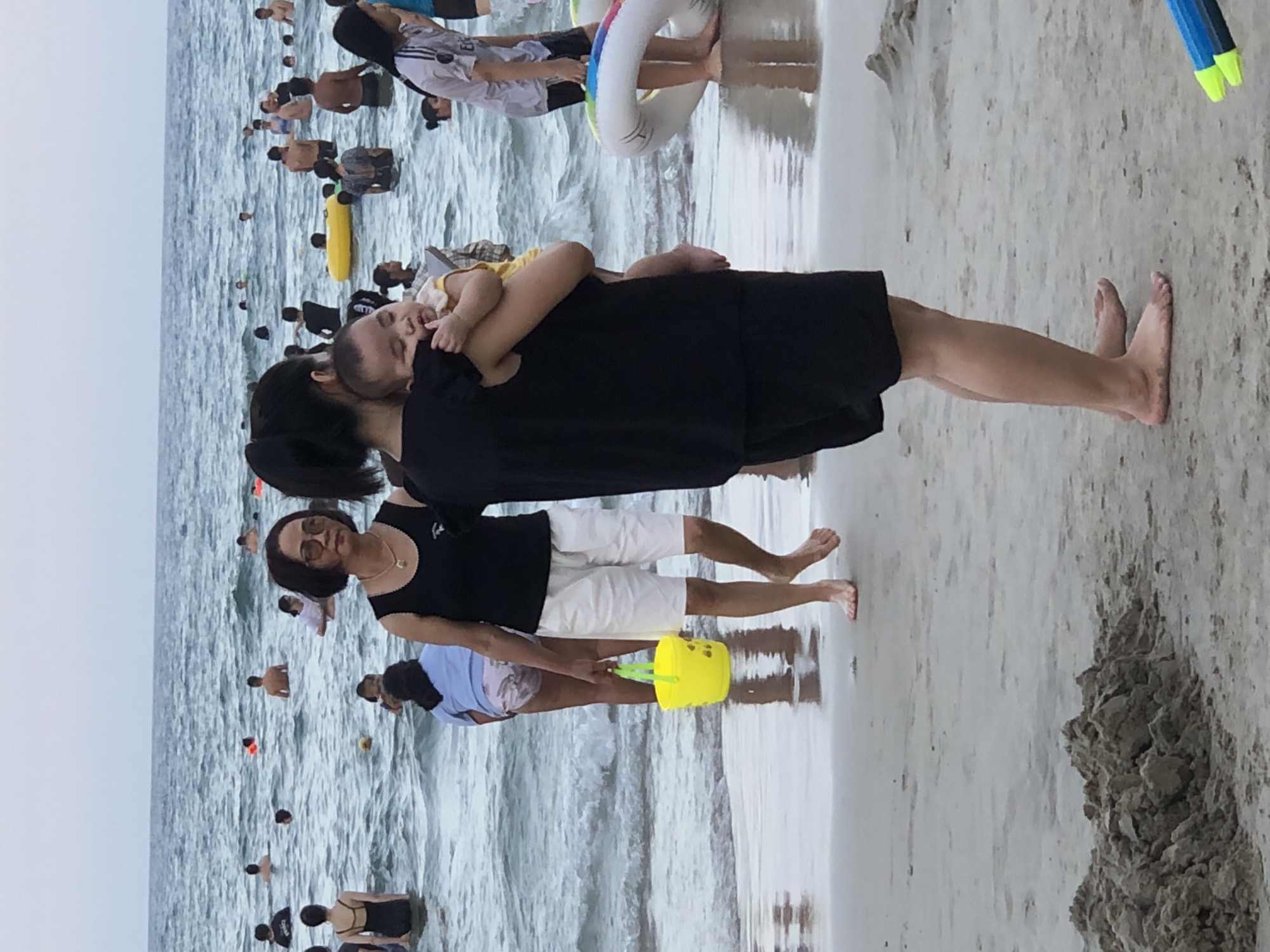 The image depicts a beach scene in Da Nang, Vietnam, where many people are enjoying the ocean. In the foreground, a woman is holding a baby, facing away from the camera. To her left, another woman, wearing a black top and white shorts, is holding a yellow bucket and looking out at the water. The ocean is filled with people swimming and playing. Some are wearing swimsuits, while others are in casual clothing. There are also inflatable toys visible in the water, suggesting recreational activities. The sand on the beach is wet, indicating the tide is close to the shore. The sky is overcast, creating a diffused light over the scene.