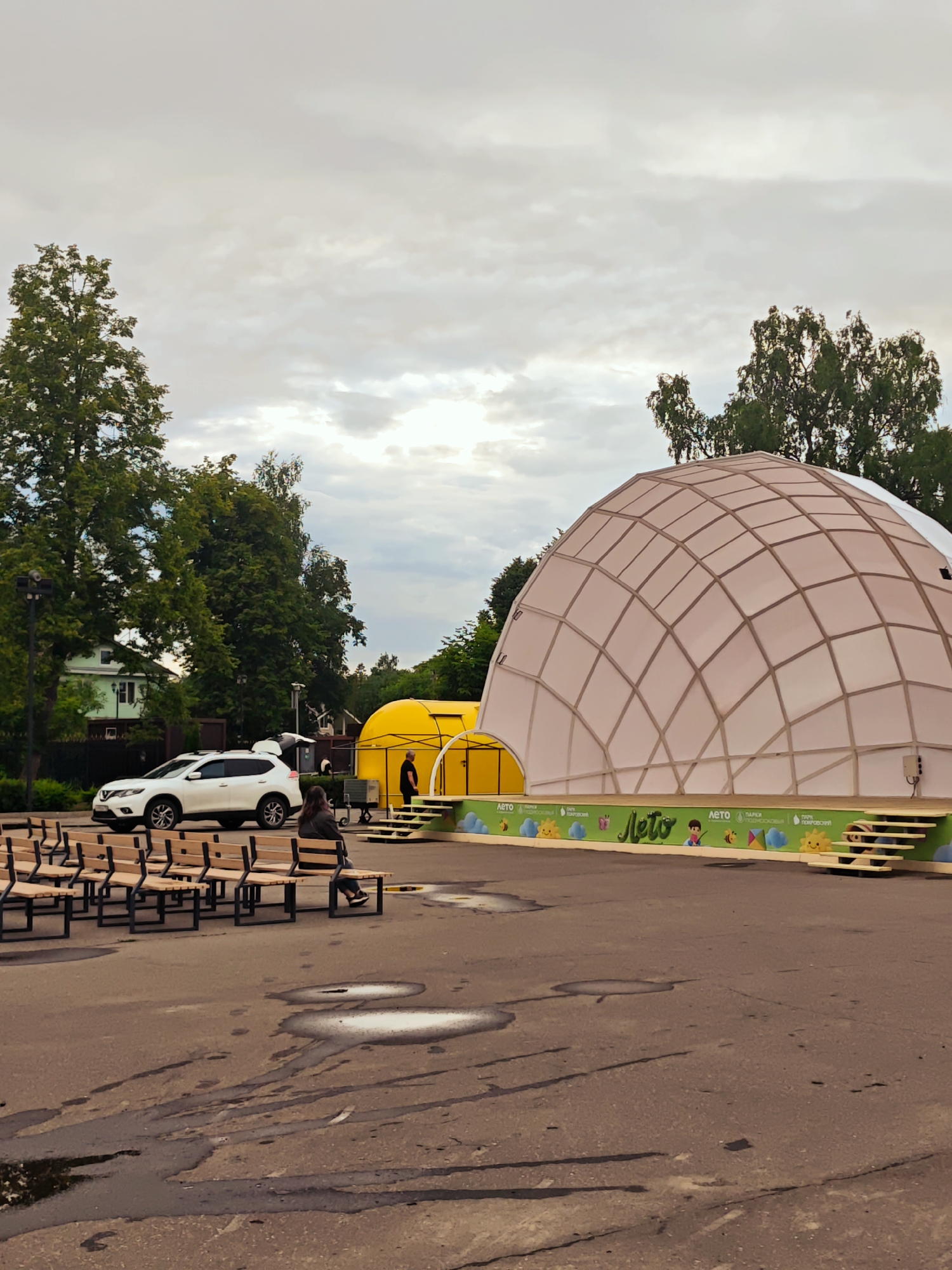 The image depicts an outdoor event space in Khotkovo, Russia. In the foreground, rows of empty wooden benches with metal frames are arranged on a paved area. Further back, a white SUV with its trunk open is parked. To the right of the SUV is a large, translucent dome structure with a geodesic frame, suggesting it might be a temporary pavilion or stage cover. Adjacent to the dome is a yellow, tent-like structure. A person is seated on one of the benches facing away from the camera, and another person stands near the yellow structure. The background features mature trees and a building, and the sky is overcast with clouds. The ground in front of the benches shows several puddles. A decorative banner with text and cartoon-like illustrations is visible along the front of the stage area.
