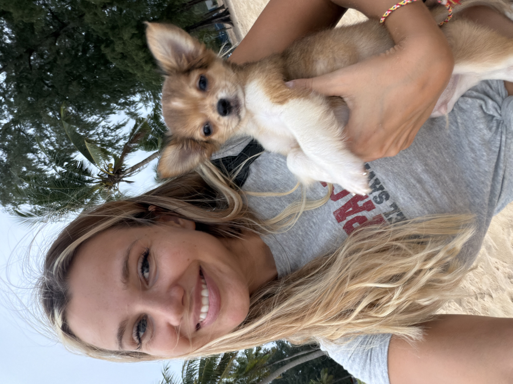 A young woman with blonde hair is holding a small tan and white puppy on a beach in Choeng Thale, Thailand. The woman is wearing a grey t-shirt with red lettering. The puppy has large ears and is looking at the camera. In the background, there are palm trees and a cloudy sky.