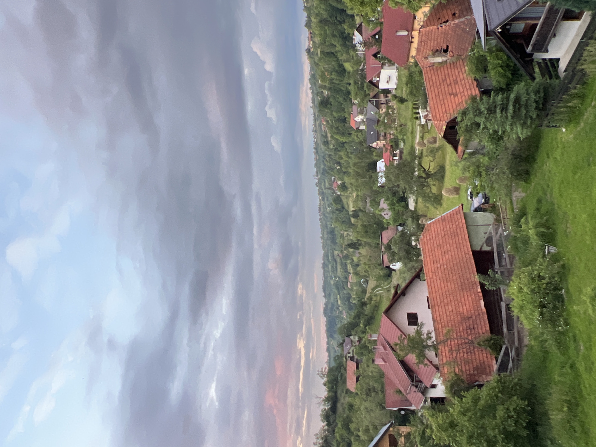 The image shows a rural village nestled in rolling hills under a cloudy sky. The foreground is dominated by green grass and lush vegetation, with several houses featuring red-tiled roofs scattered throughout. In the midground, more houses and trees create a dense, wooded landscape. The background features further hills covered in trees, with a winding road visible. The sky is partly cloudy, with patches of soft pink and blue hues indicating either sunrise or sunset. The overall impression is one of a peaceful, natural setting.